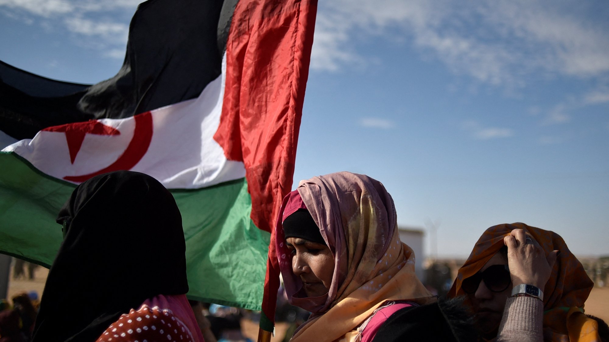 Des femmes portant une écharpe colorée et une grande bannière. Ciel dégagé en arrière-plan.