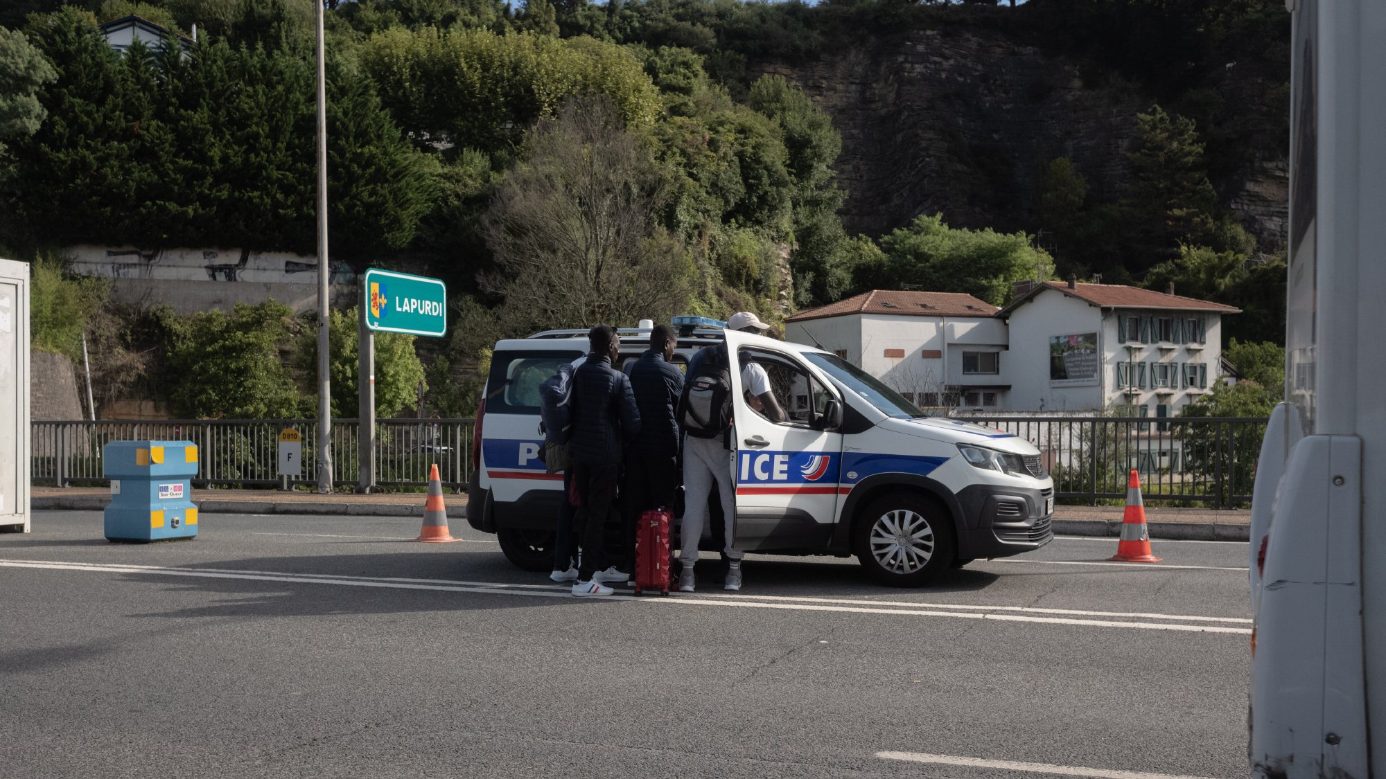 The image shows a scene on a road where a police vehicle is stopped. A few individuals are standing near the back of the police car, which is marked with the word "Police." The setting appears to be a highway or a major road, with some road cones placed nearby. In the background, there are trees and some buildings visible, suggesting an urban or suburban area. The atmosphere seems to indicate a police check or an interaction taking place.