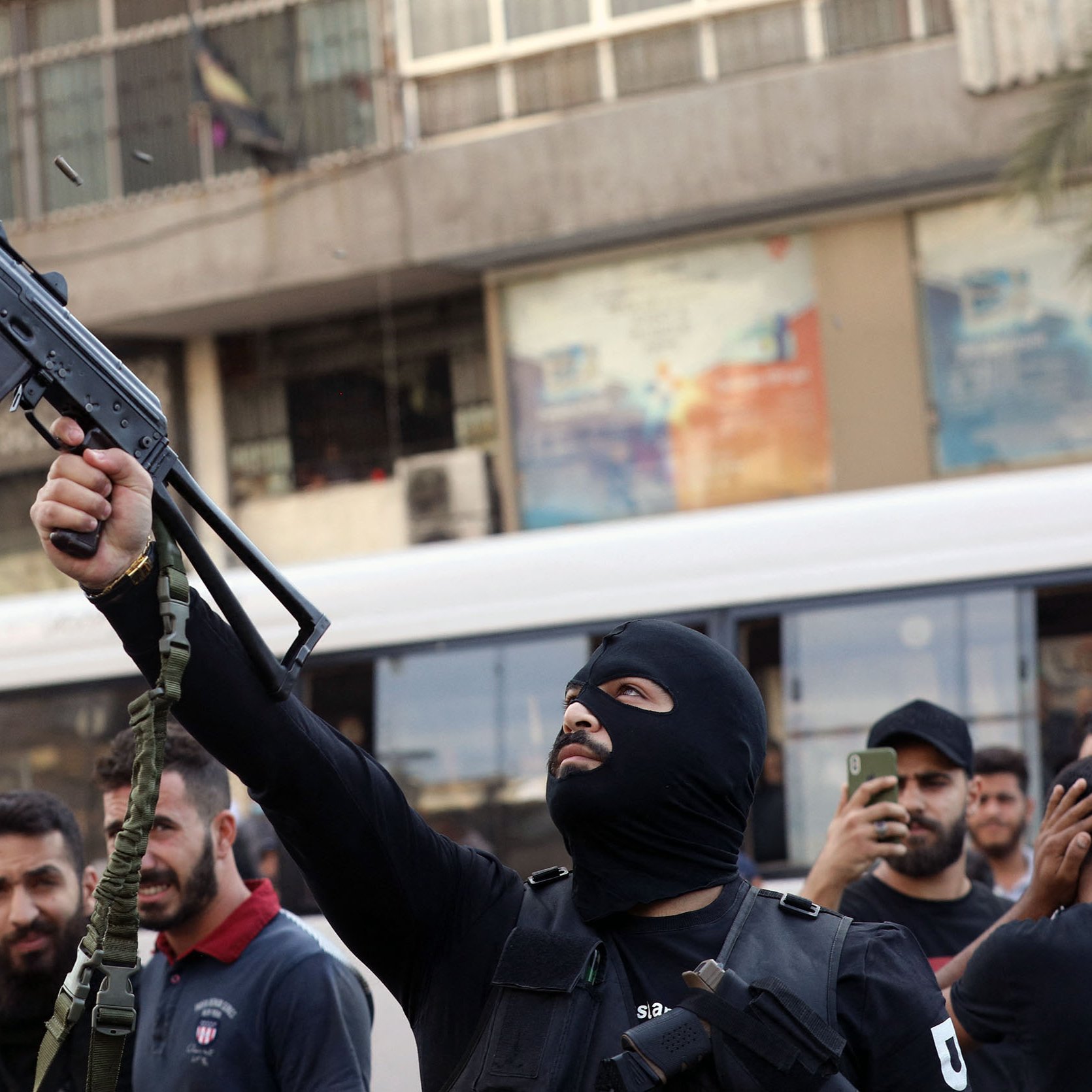 L'immagine mostra un uomo mascherato che brandisce un fucile automatico in una strada affollata. Si possono vedere altre persone intorno a lui, alcune delle quali sembrano sostenere o osservare l'azione. L'ambiente circostante è urbanizzato, con edifici e un autobus che appare in lontananza. L'atmosfera sembra carica di tensione, suggerendo un contesto di protesta o conflitto.