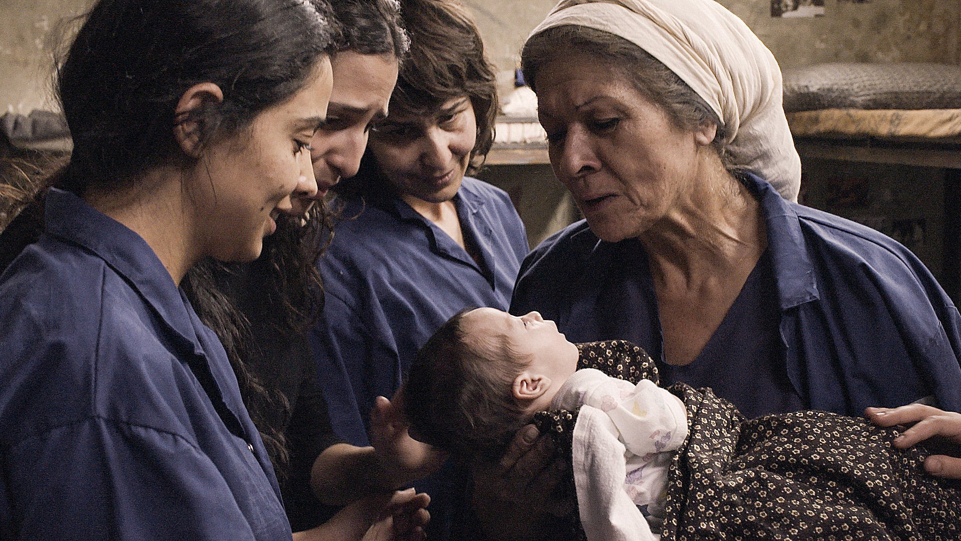 L'image montre un moment touchant où une femme âgée tient un bébé dans ses bras, partageant une interaction affectueuse. Autour d'elle, trois jeunes femmes observent et semblent émerveillées par ce moment. Elles portent toutes des vêtements similaires, de couleur bleue, et l'environnement semble simple et chaleureux. L'atmosphère dégage une sensation de tendresse et de solidarité entre les générations.