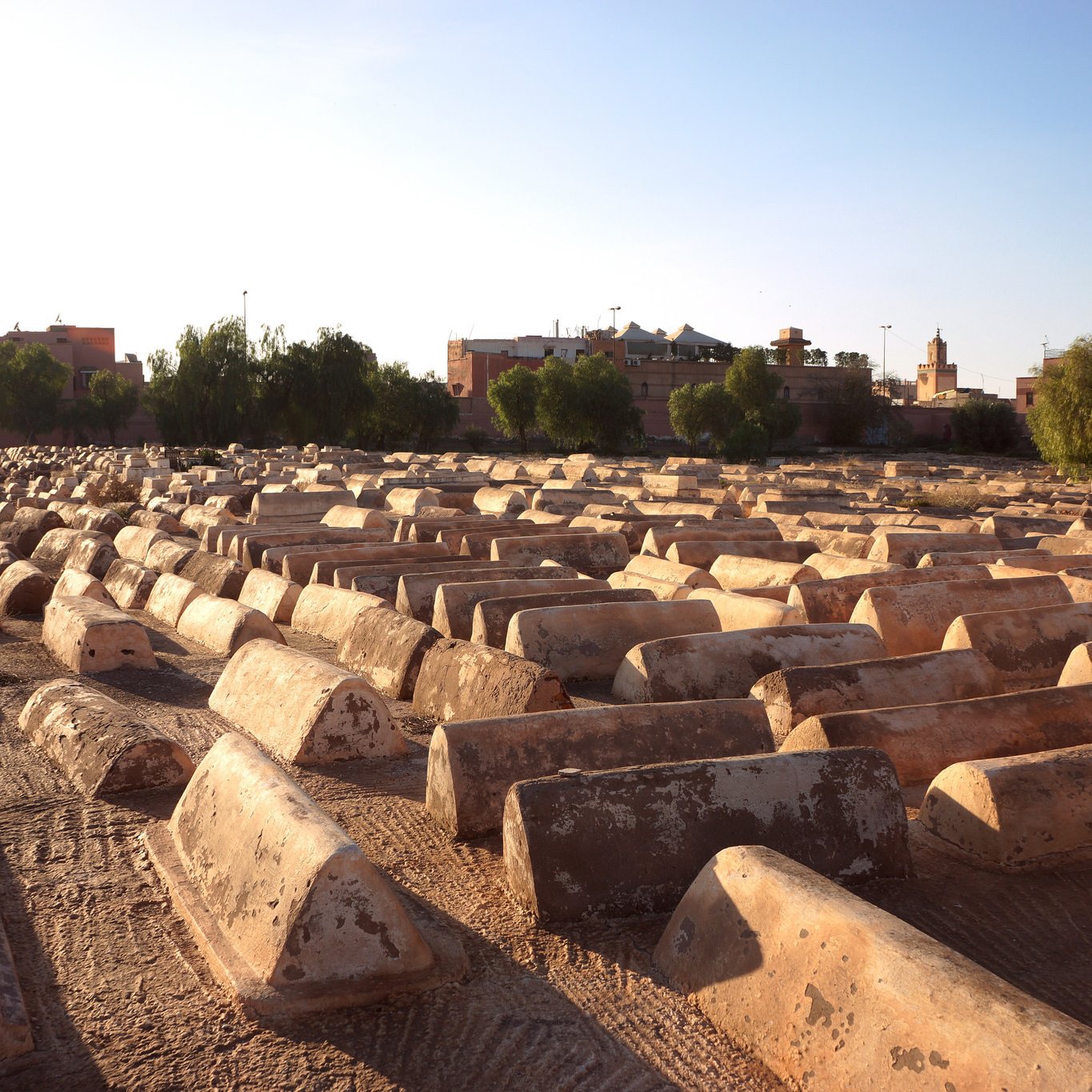 L'image montre un cimetière où l'on peut voir des tombes en forme de tombeaux, étalées sur un terrain sec. Les structures sont de couleur beige ou terre cuite et semblent être en béton, avec une surface usée par le temps. En arrière-plan, on aperçoit des bâtiments et des arbres, indiquant que le cimetière est situé près d'une ville. La lumière du soleil crée une atmosphère calme et paisible, tandis que le ciel est dégagé, laissant entrevoir un beau jour.