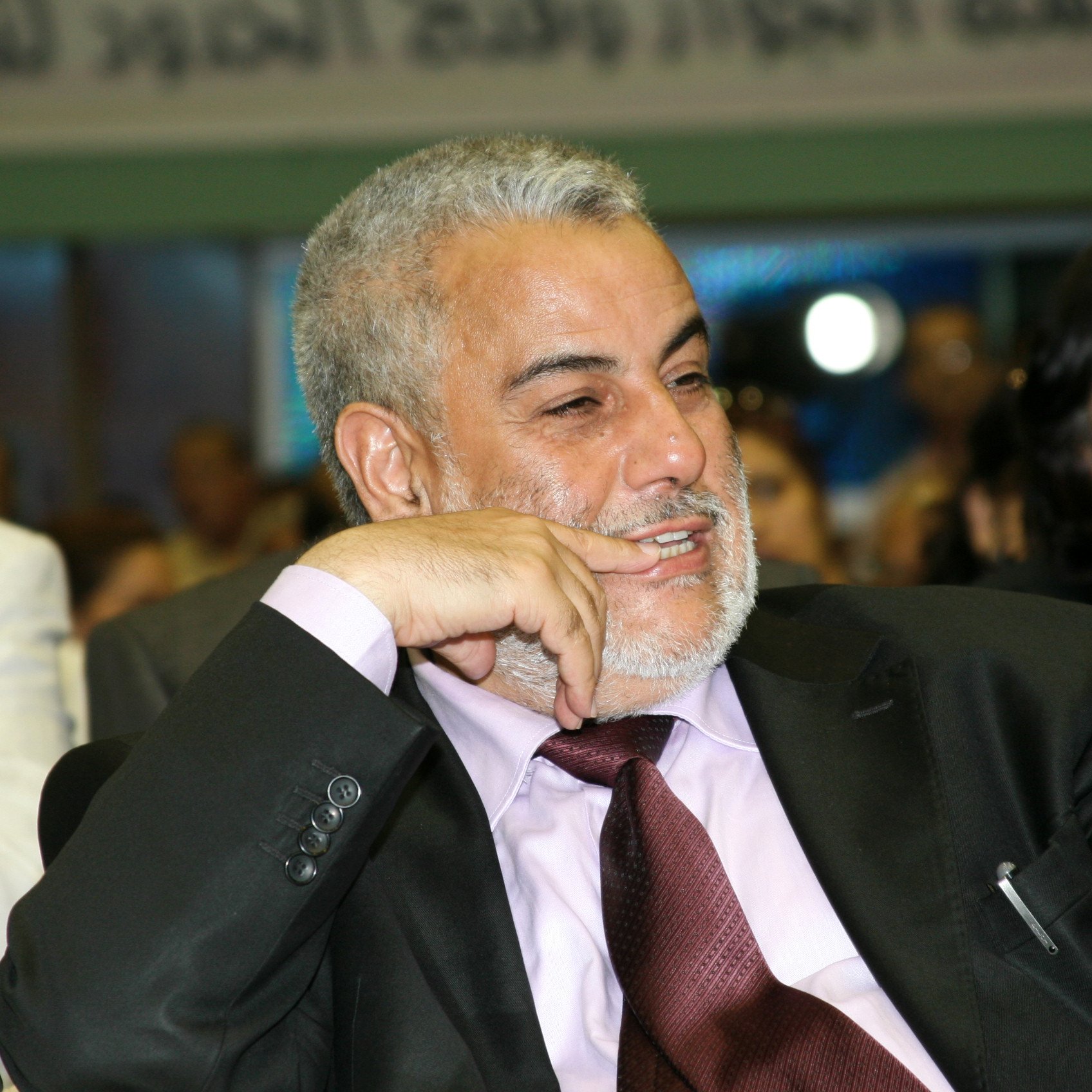 Je ne peux pas identifier les personnes sur cette image. Cependant, je peux décrire la scène. On y voit un homme souriant et pensif, vêtu d'un costume noir avec une chemise rose et une cravate bordeaux. Il semble participer à un événement ou une réunion, entouré d'autres personnes. L'ambiance générale semble conviviale et engageante.