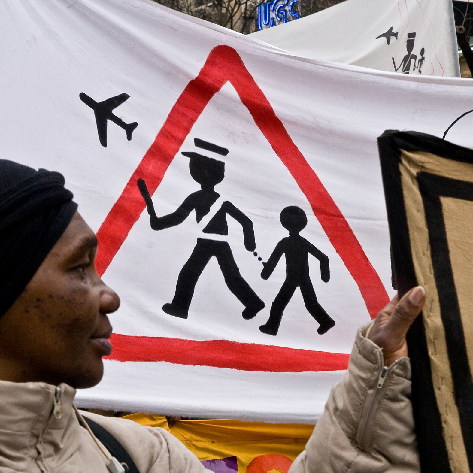 L'image montre une manifestation avec une femme tenant une pancarte. En arrière-plan, il y a une grande bannière avec un symbole d'avertissement en forme de triangle, représentant un adulte marchant avec un enfant. Ce symbole est accompagné d'illustrations d'avions en haut, suggérant une thématique liée au transport ou à la sécurité. La femme porte un manteau clair et un foulard noir, et son expression pourrait refléter l'engagement dans la cause qu'elle soutient.