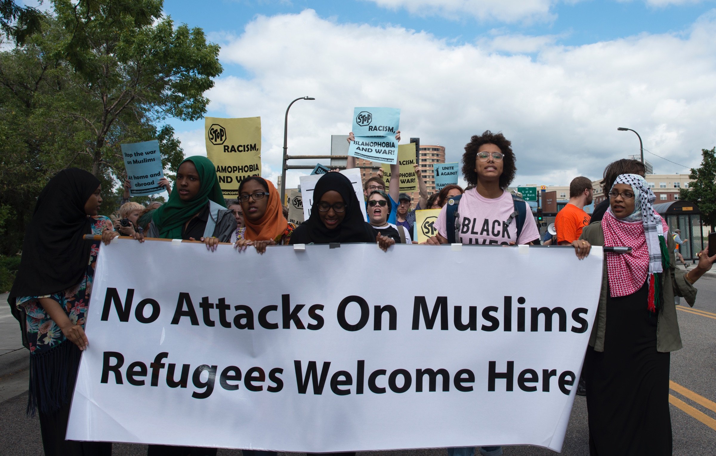 L'image montre un groupe de manifestants en train de marcher dans une rue. Ils portent des pancartes avec des messages tels que "No Attacks On Muslims" et "Refugees Welcome Here". Les participants, majoritairement de culture musulmane, affichent des expressions déterminées. Le ciel est partiellement nuageux, et on peut voir des bâtiments en arrière-plan. L'atmosphère semble engager et militante, soulignant des thèmes de solidarité et de droits humains.