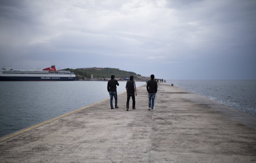 L'image montre trois personnes marchant sur un quai. Le ciel est nuageux, et l'atmosphère est calme. À l'arrière-plan, on peut voir un ferry amarré et une petite île. Le quai est en béton, et l'eau semble calme, reflétant les nuages. Les silhouettes des gens ajoutent une touche de vie à cette scène paisible.