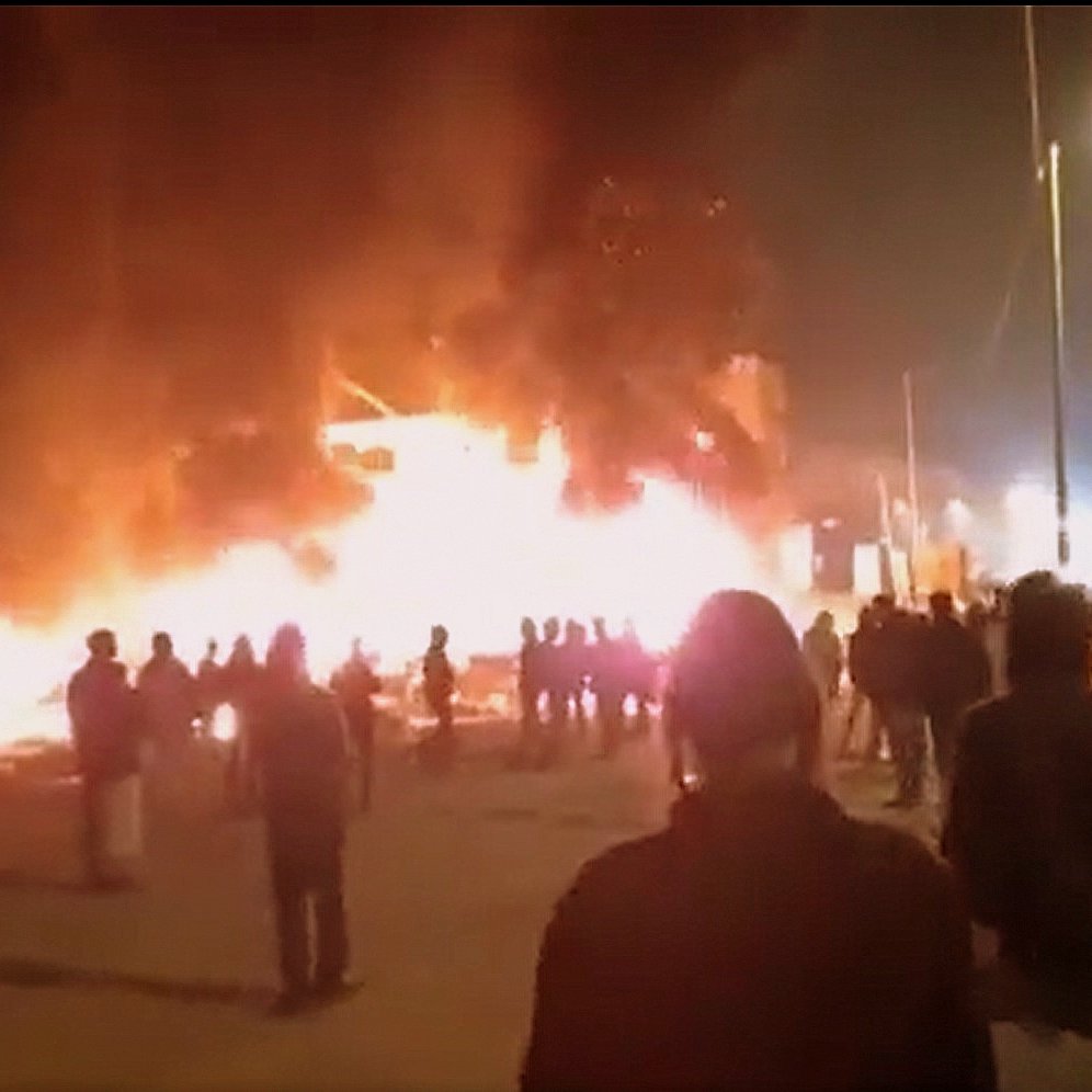 L'image montre une scène nocturne où un bâtiment est enflammé, créant une grande colonne de feu et de fumée. Des personnes se tiennent au premier plan, observant la situation. On peut noter une ambiance de tension, avec des lumières des bâtiments environnants qui ajoutent à l'atmosphère dramatique. Les silhouettes des personnes sont découpées contre la lumière des flammes.