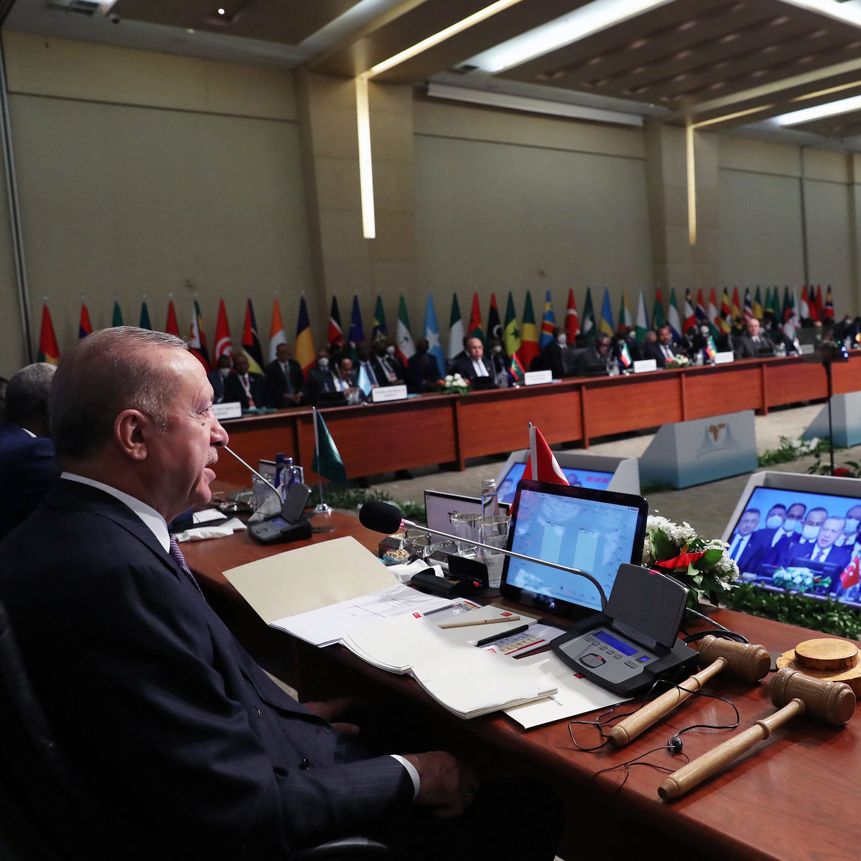 تظهر الصورة اجتماعًا رسميًا يحمل طابعًا دوليًا، حيث يتواجد عدد من القادة أو المسؤولين الجالسين على طاولة مستديرة. في مقدمة الصورة، يظهر رجل يجلس أمام طاولة، ويرتدي بدلة، بينما خلفه مجموعة من الأعلام الوطنية للدول المختلفة. تُظهر الشاشة خلفهم مجموعة من الأشخاص، ويبدو أن الاجتماع يناقش قضايا مهمة. يُحيط بالطاولة بعض الزهور والزينة، مما يضفي طابعًا رسميًا على الحدث.