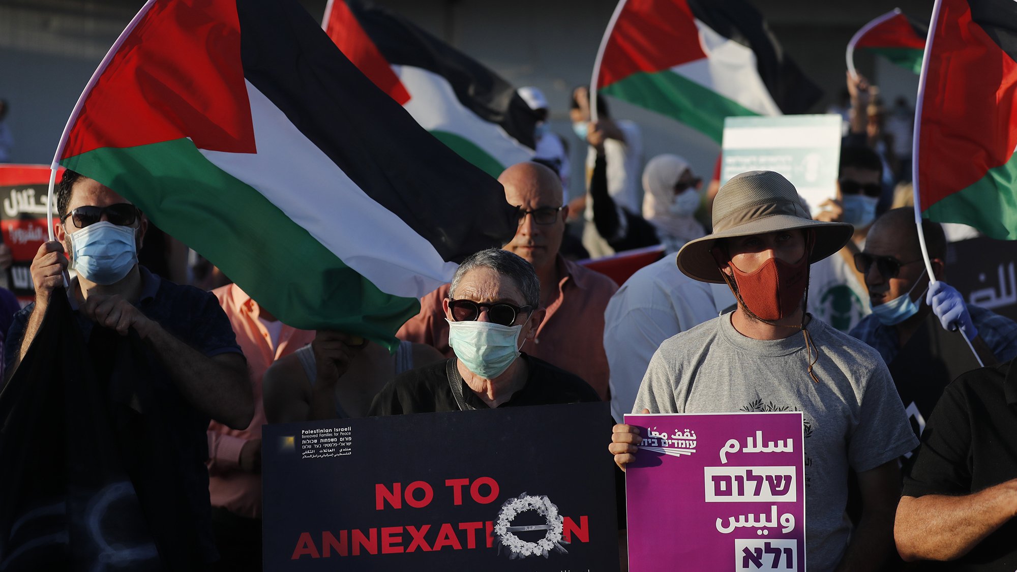 در این تصویر، گروهی از مردم در حال برگزاری یک تظاهرات هستند. آنها پرچم‌های فلسطینی را در دست دارند و تعدادی از آنها تابلوهای اعتراضی حمل می‌کنند. نوشته‌های روی تابلوها شامل عباراتی مانند "نه به الحاق" و "بیایید خانواده‌های بیشتری را اضافه نکنیم" به چشم می‌خورد. همچنین، برخی از تظاهرکنندگان ماسک به صورت دارند. فضای تصویر به وضوح نشان‌دهنده اعتراض و حمایت از حقوق فلسطینیان است.