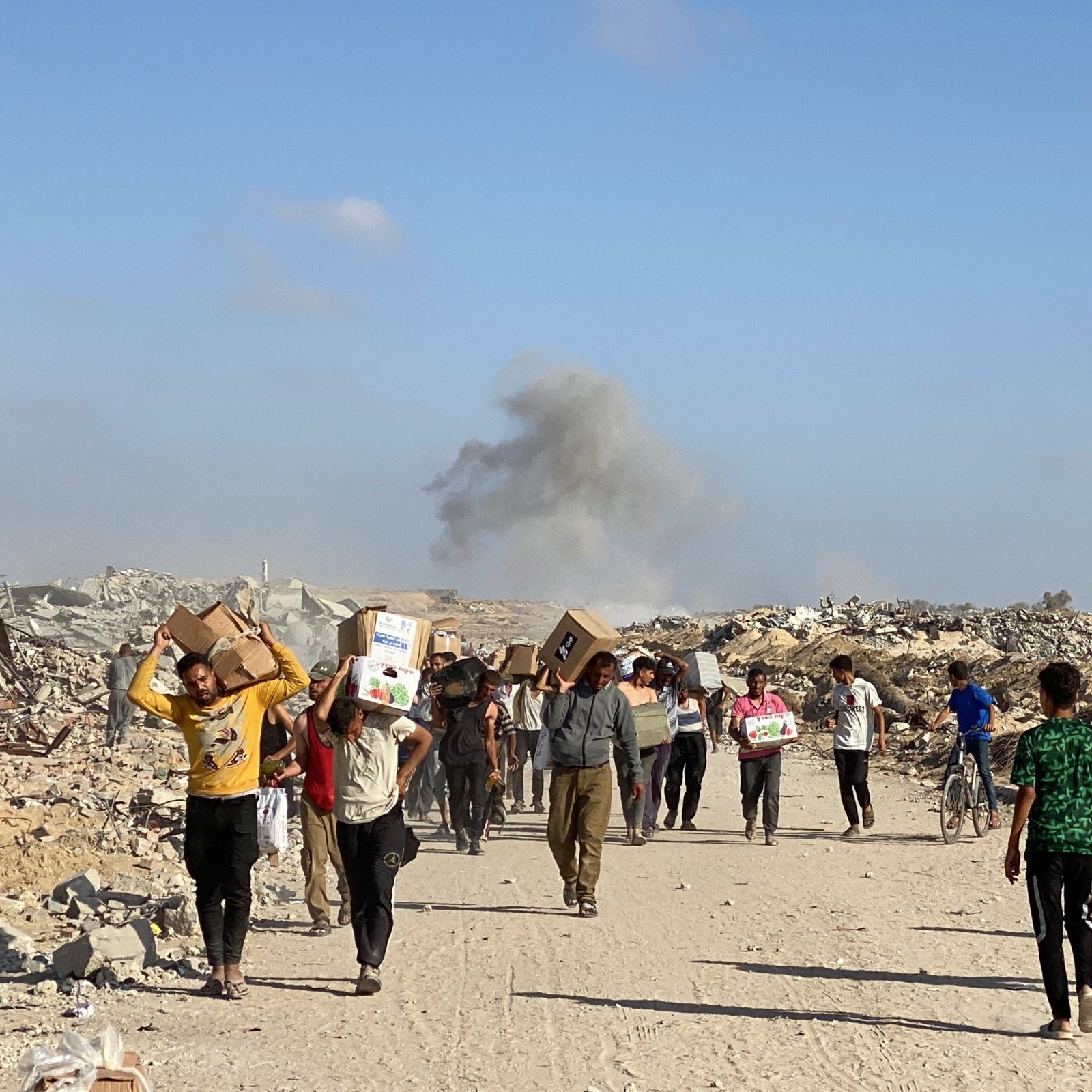 Des personnes marchent dans des décombres, portant des colis, avec de la fumée au loin.