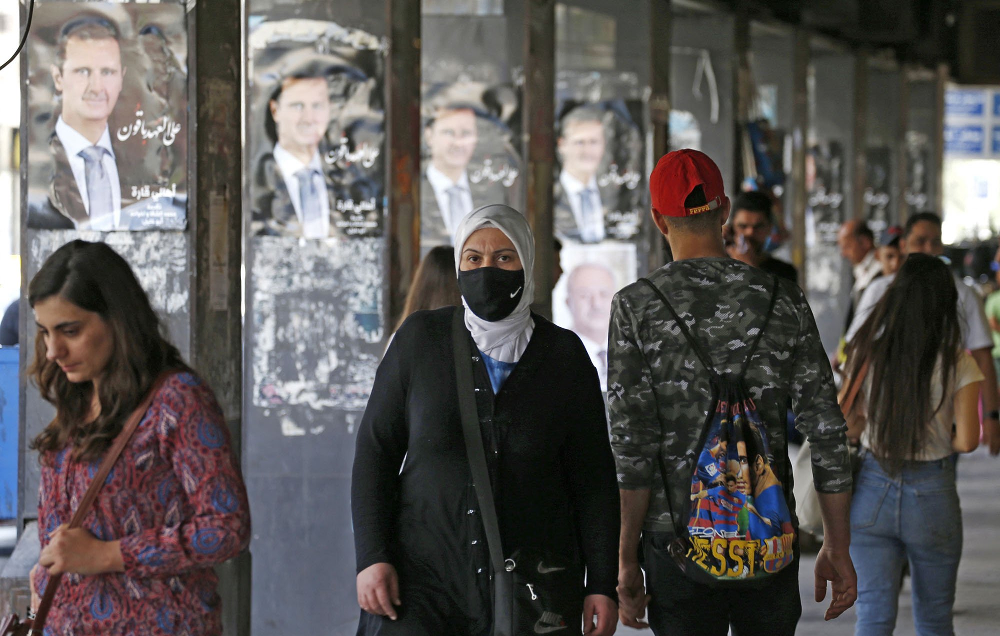 L'image montre une rue animée avec des personnes marchant sous un abri. En arrière-plan, on aperçoit des affiches de propagande, probablement de l'homme politique en question. Plusieurs personnes, dont une femme portant un hijab et un masque, se déplacent le long du trottoir. L'atmosphère semble dynamique et quelque peu urbaine, avec des jeunes et des adultes interagissant dans cet espace public.