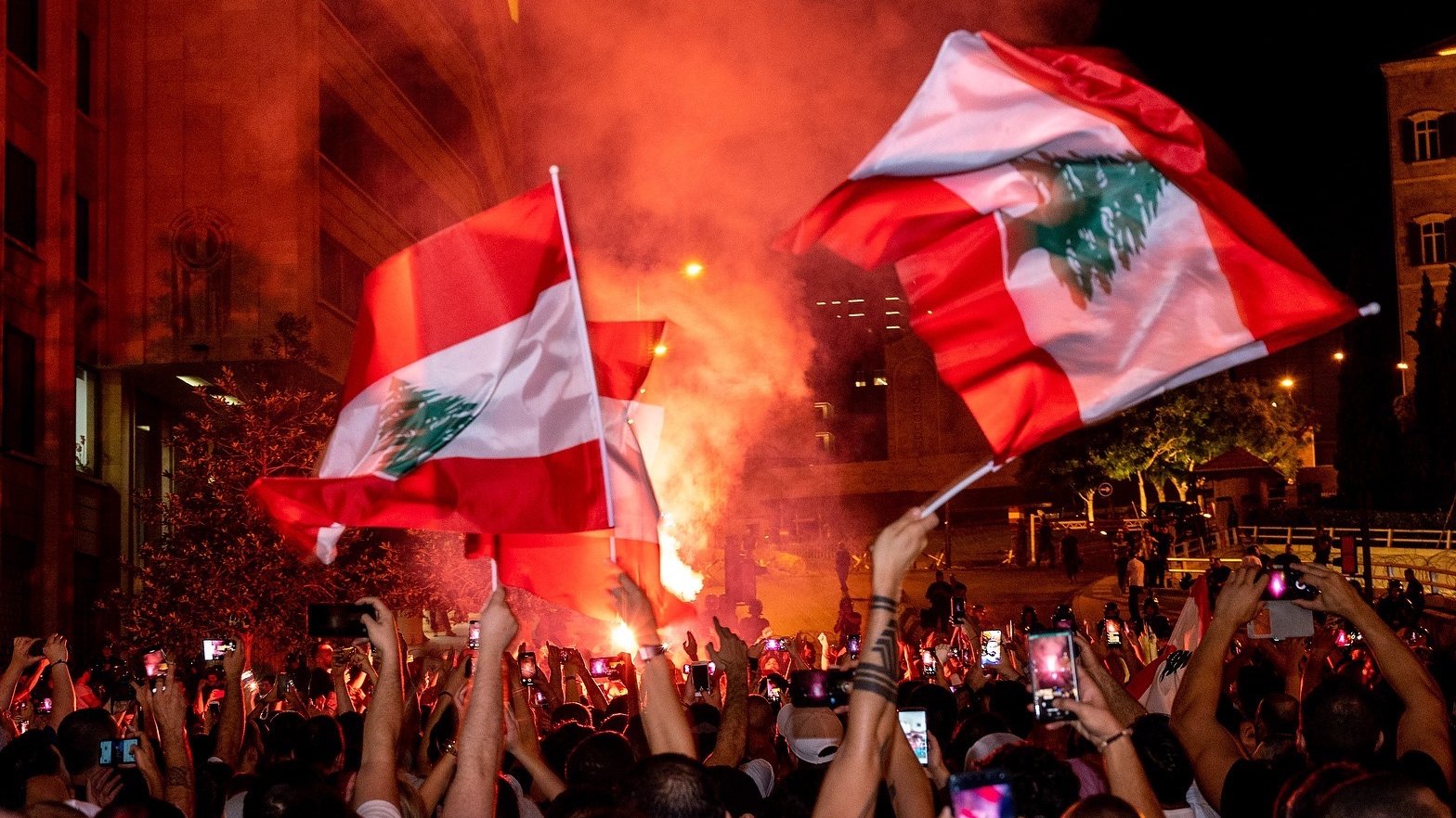 The image depicts a large gathering of people celebrating, likely in an urban setting. Many individuals are holding flags of Lebanon, which feature a red and white design with a green cedar tree. The atmosphere appears vibrant and energetic, with colorful smoke and lights adding to the scene. People are also raising their phones to capture the moment, suggesting a significant event or celebration taking place.