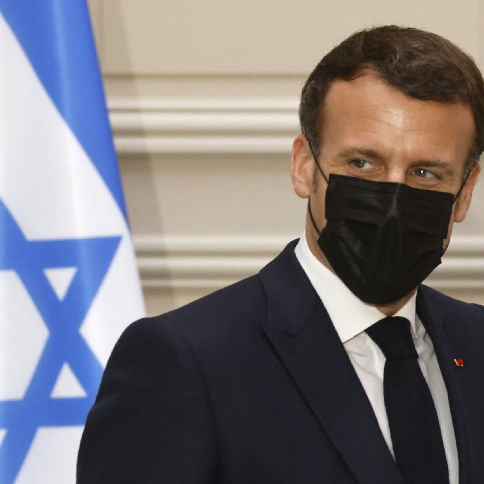 الصورة تظهر رجلًا يرتدي بدلة رسمية وقناعًا أسود. خلفه يوجد علم يحمل نجمة داود. يبدو الرجل في وضعية جانبية، حيث يلتفت نحو الجانب. تعكس الإضاءة في الصورة أجواء رسمية. لا يمكنني تحديد من هو هذا الشخص.