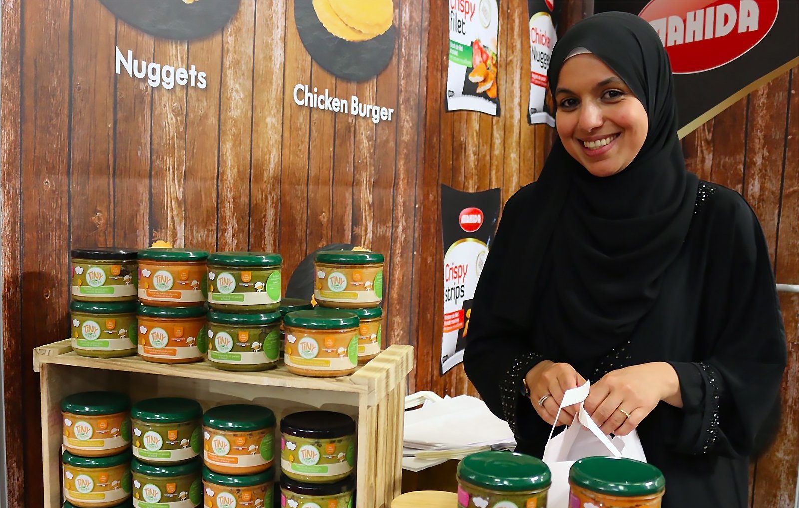 L'image montre une femme portant un hijab, souriant tout en se tenant près d'un stand de produits alimentaires. À côté d'elle, il y a une étagère en bois remplie de pots de produits, probablement des sauces ou des condiments, étiquetés avec différentes saveurs. L'arrière-plan est en bois, et on peut voir d'autres affiches promotionnelles. L'ambiance semble conviviale, et la femme semble enthousiaste à propos des produits exposés.