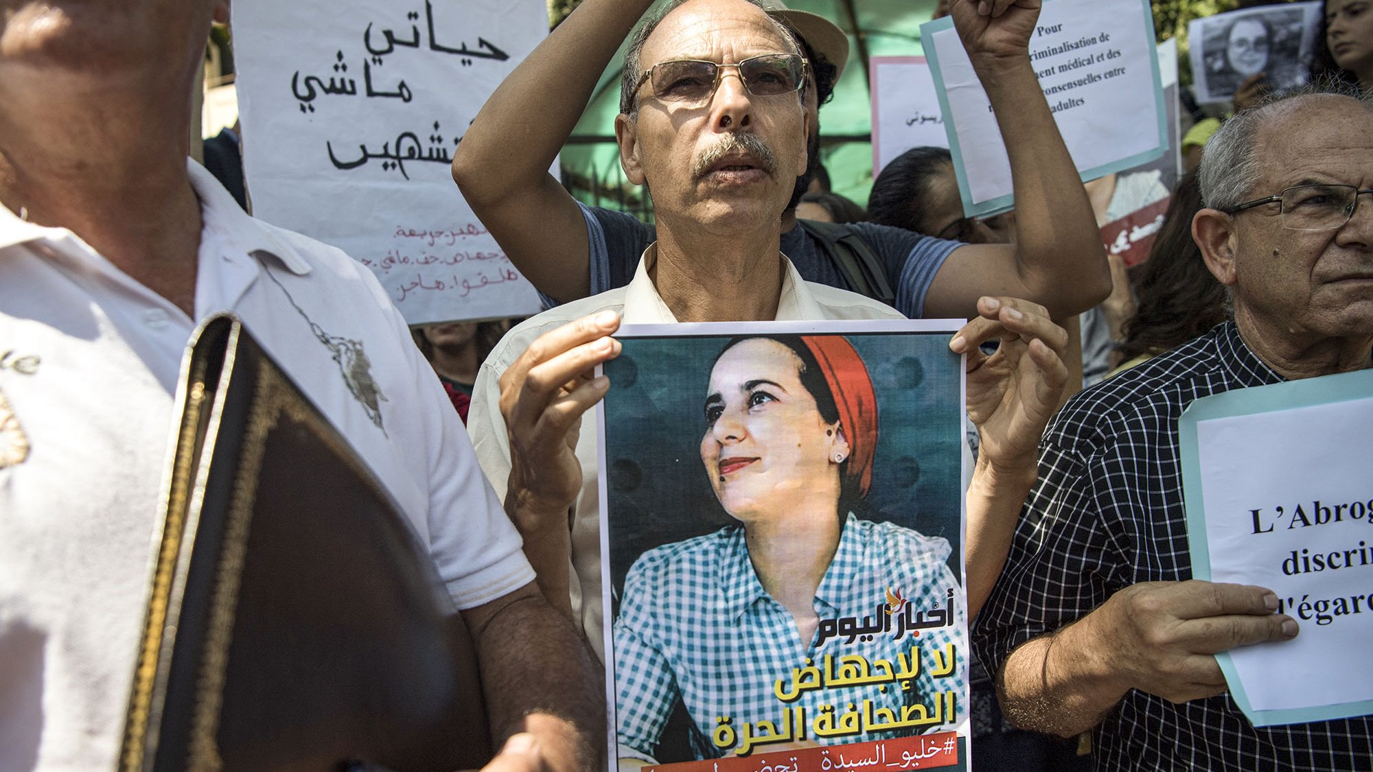 L'image montre un groupe de personnes rassemblées pour une manifestation. Au premier plan, un homme tient une affiche qui représente une femme avec un sourire, probablement une figure symbolique pour le mouvement. Les participants, majoritairement des adultes, tiennent également des pancartes avec des slogans en arabe et en français, indiquant leur lutte ou leur soutien à une cause, sans doute liée à des droits ou à une injustice. L'atmosphère semble déterminée et engagée.