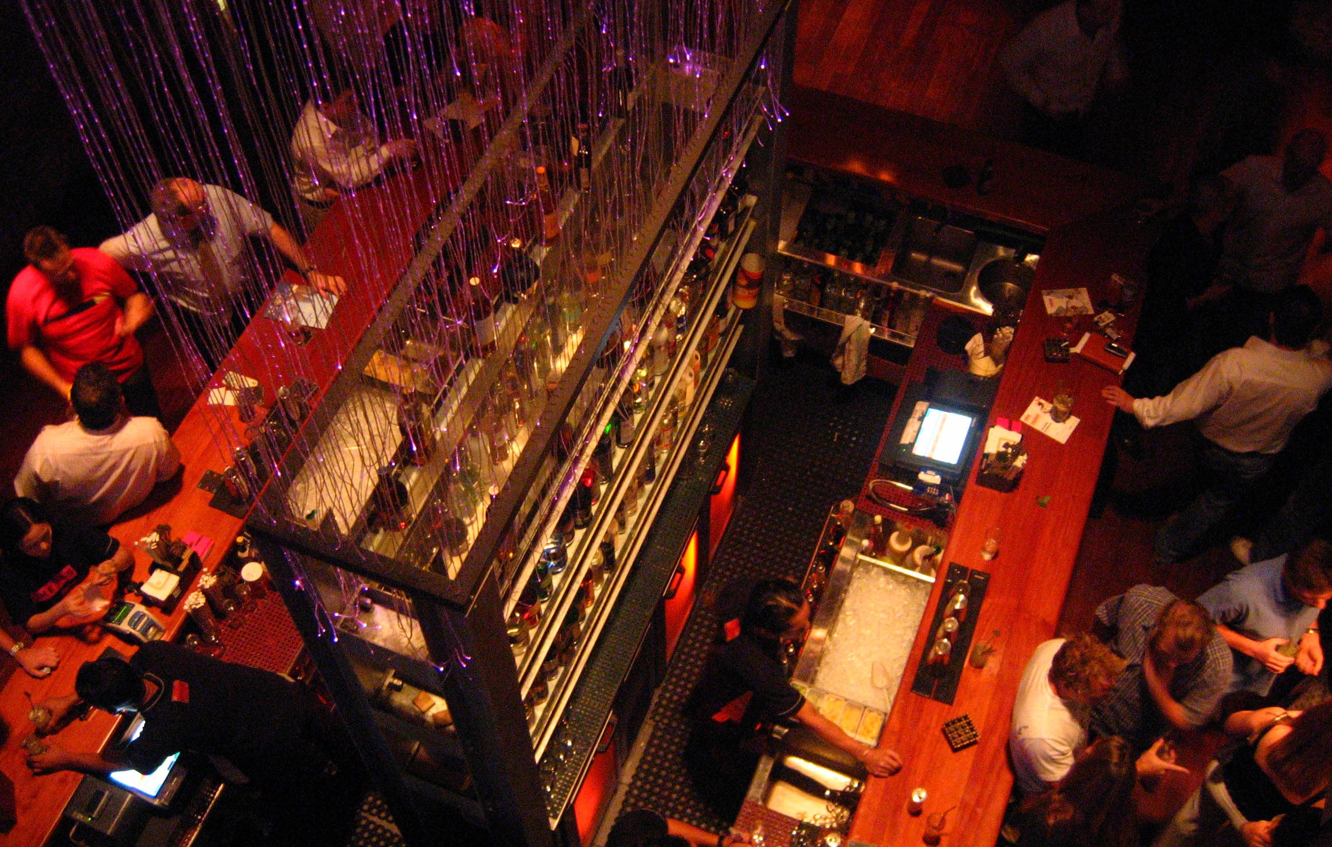 L'image montre une vue aérienne d'un bar animé. On peut voir une grande table de bar en bois clair, avec de nombreuses personnes stationnées autour, se parlant et déguster des boissons. Au dessus, il y a des chaînes de lumières, créant une ambiance festive. Les étagères à bar sont bien garnies de bouteilles colorées, et on distingue des serveurs s'affairant derrière le comptoir. L'atmosphère semble conviviale et dynamique, avec une décoration moderne.