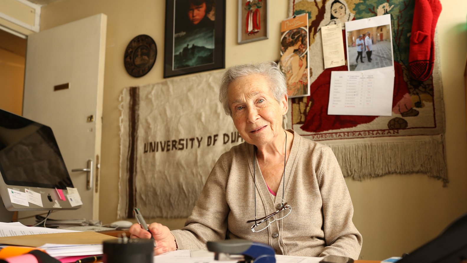 L'image montre une femme âgée assise à un bureau. Elle porte un pull beige et a des lunettes qui reposent sur son nez. Son sourire est chaleureux et accueillant. Le bureau est en désordre avec des papiers éparpillés, un agrafeuse et un verre. En arrière-plan, on aperçoit divers objets accrochés au mur, y compris un calendrier et des photos. On peut aussi distinguer une toile ou un tissu qui mentionne "UNIVERSITY OF DUBAI". L'atmosphère est conviviale et studieuse.