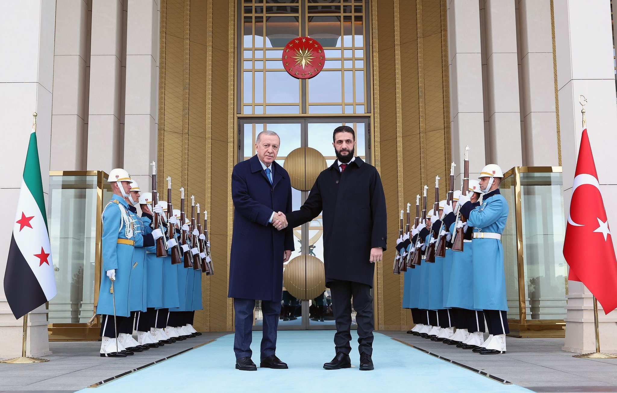 L'image montre deux hommes se serrant la main devant un bâtiment officiel. Ils sont entourés de gardes en uniforme bleu, tenant des fusils, et un grand symbole turc est visible derrière eux. La scène dégage une atmosphère solennelle.