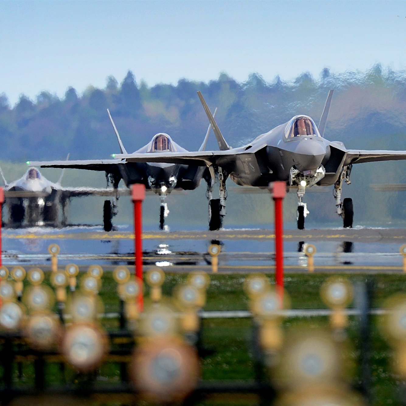 تظهر في الصورة ثلاث طائرات مقاتلة من طراز F-35 تتواجد على مدرج مطار. الطائرات تبدو وكأنها جاهزة للإقلاع، حيث تبرز من الخلف ملامحها الحديثة وتصميمها الأنيق. الأرضية محاطة بمجموعة من الحواجز أو الأعمدة، في حين أن المناظر الطبيعية في الخلفية تظهر أشجارًا ومساحات خضراء تحت سماء زرقاء. تُعطي الصورة انطباعًا عن القوة والتقنية المتقدمة في مجال الطيران العسكري.