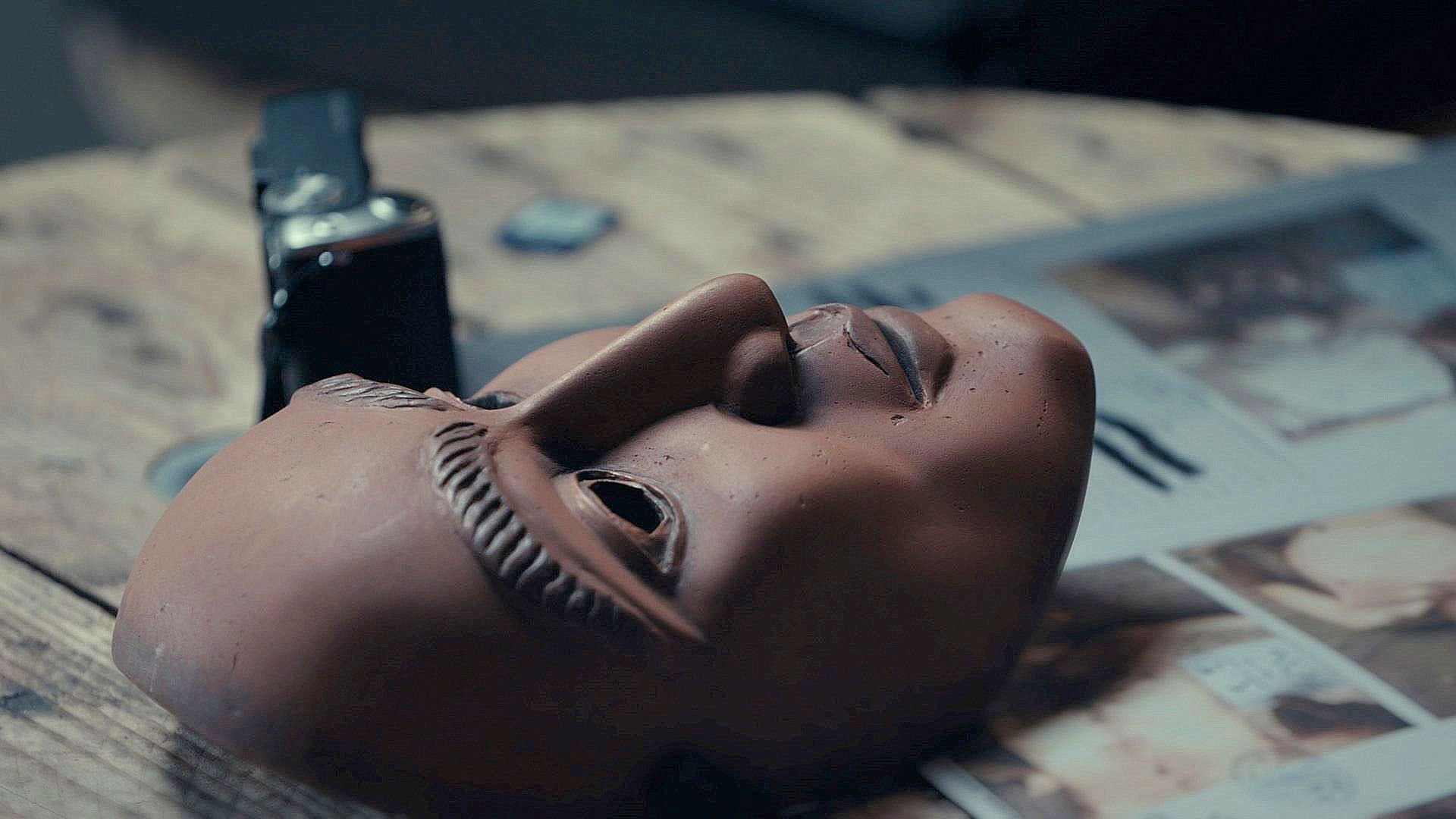 L'image montre un masque en terre cuite posé sur une surface en bois. Le masque a des traits détaillés, avec une expression neutre et des sourcils sculptés. À côté du masque, on voit une montre ou un objet similaire, ainsi qu'un magazine ouvert avec des illustrations. L'éclairage est doux, créant une ambiance contemplative autour de ces objets.