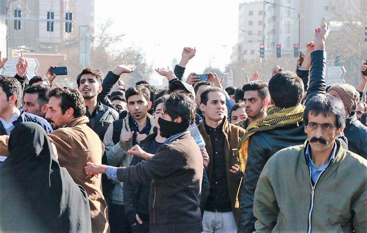 L'image montre une foule de personnes rassemblées dans une rue. Les individus semblent engagés dans une forme de manifestation ou de rassemblement collectif. Certains lèvent les mains ou filment avec des téléphones, tandis que d'autres ont des expressions préoccupées ou déterminées. On peut voir une diversité d'habillements, y compris des vêtements traditionnels, et un mélange de genres parmi les participants. L'ambiance générale semble être celle de l'enthousiasme ou de la passion pour une cause commune.