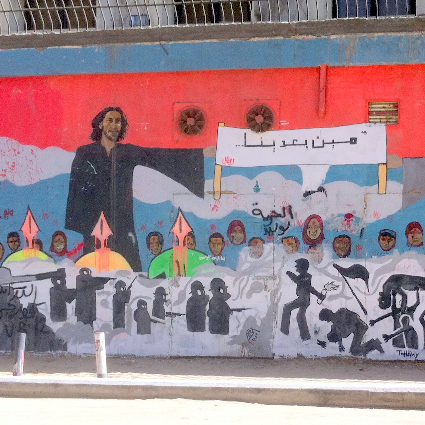 L'image montre une grande fresque murale qui illustre des figures humaines et des symboles de résistance. À gauche, on voit une personne avec de longs cheveux, et à droite, un homme âgé portant un turban. Entre eux, il y a une inscription dans une langue arabe. Le mural est riche en détails, avec des silhouettes de personnes et des flèches, suggérant des thèmes de lutte et d'espoir. Le fond est principalement rouge, créant un contraste fort avec les personnages et les éléments représentés.