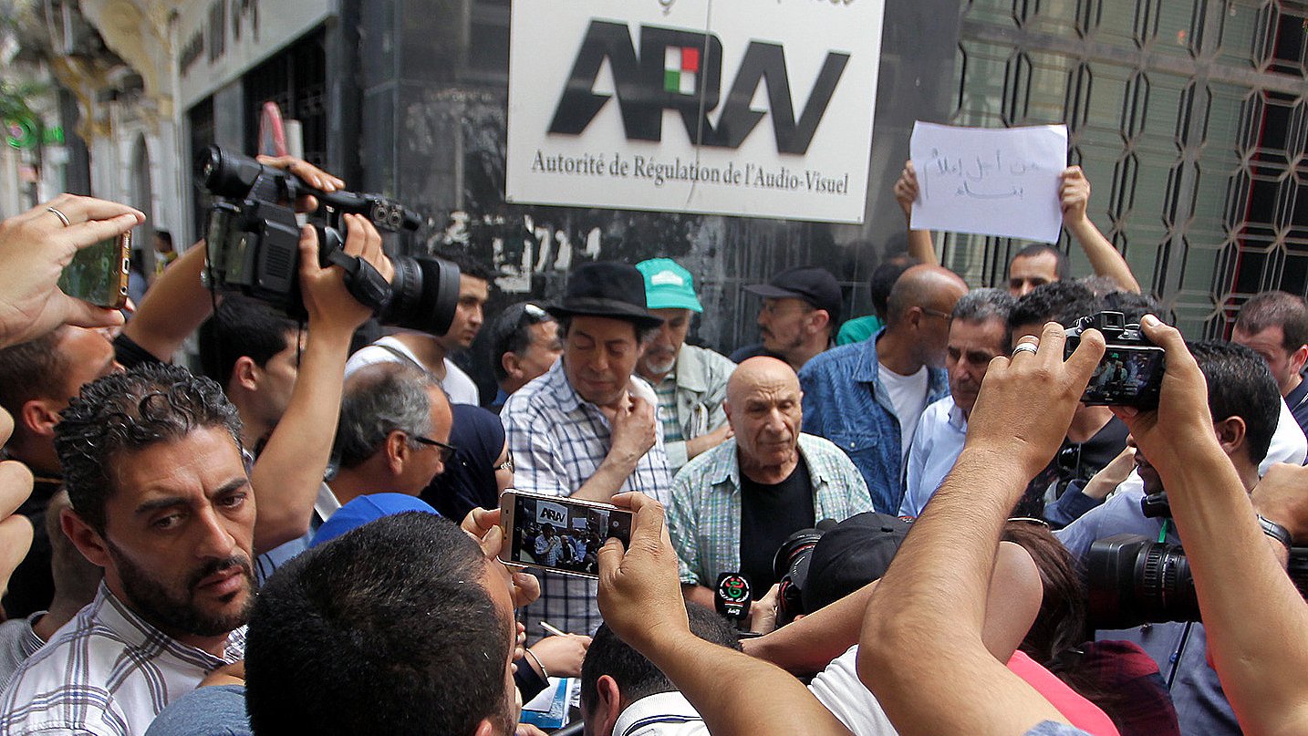 L'image montre un groupe de personnes rassemblées autour d'un homme au premier plan, visiblement en pleine discussion ou confrontation. Plusieurs caméras et téléphones portables sont levés, capturant l'événement. En arrière-plan, on peut voir une bannière avec le mot "ARAV" qui fait référence à l'Autorité de Régulation de l'Audiovisuel. La scène semble être un moment de protestation ou de manifestation, avec des expressions variées sur les visages des participants.