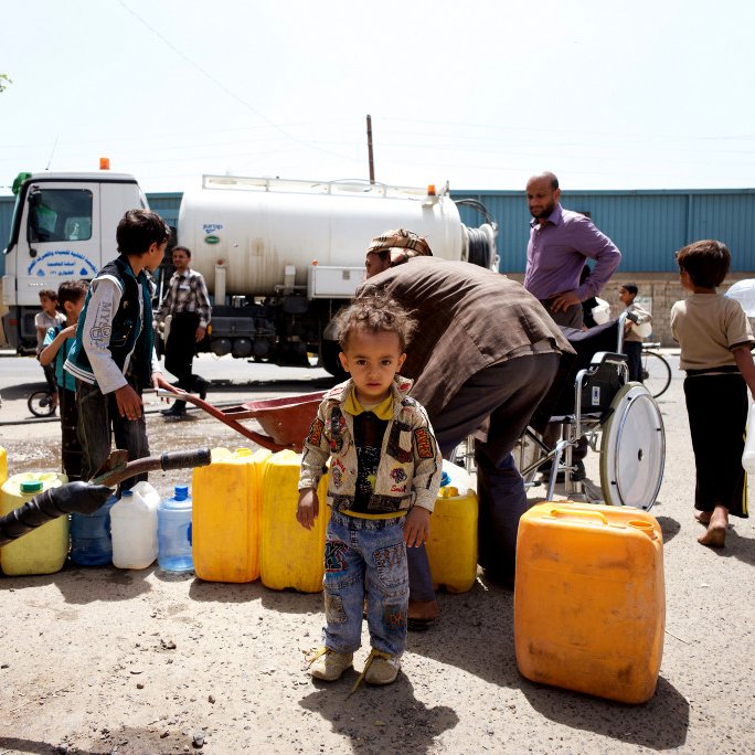 L'image montre une scène animée dans une zone urbaine, où plusieurs personnes sont rassemblées autour d'un camion-citerne. Des adultes et des enfants transportent des récipients pour collecter de l'eau. On remarque un jeune enfant au premier plan, portant une veste colorée, se tenant debout parmi d'autres personnes qui s'affairent. En arrière-plan, on aperçoit un homme en fauteuil roulant qui semble aider. Les couleurs des récipients sont vives, principalement jaunes et bleus, et l'atmosphère illustre un besoin urgent d'approvisionnement en eau.