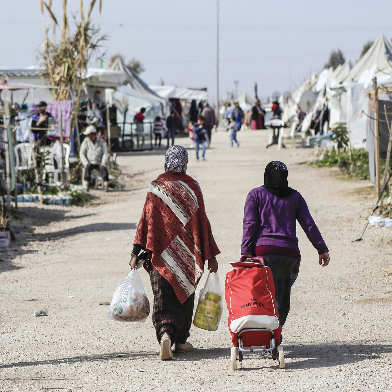 L'image montre une scène d'un camp, probablement un camp de réfugiés ou un site temporaire. Deux femmes marchent sur un chemin de terre, portant des sacs. L'une d'elles pousse une poussette. En arrière-plan, on aperçoit des tentes et des structures improvisées, avec d'autres personnes visibles dans l'environnement. L'atmosphère semble être celle d'une communauté, avec des éléments de vie quotidienne.