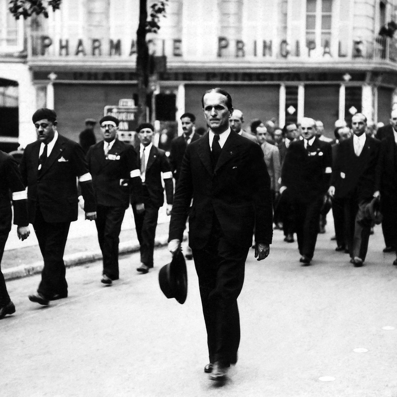 L'image montre un groupe de personnes défilant dans une rue, probablement lors d'un événement formel ou d'une commémoration. Au premier plan, un homme en costume noir porte un chapeau et marche avec assurance. Les autres participants portent aussi des costumes et certains semblent avoir des insignes ou des decorations. En arrière-plan, on peut voir un bâtiment avec l'enseigne "PHARMACIE PRINCIPALE". L'atmosphère générale de la scène semble sérieuse et respectueuse.