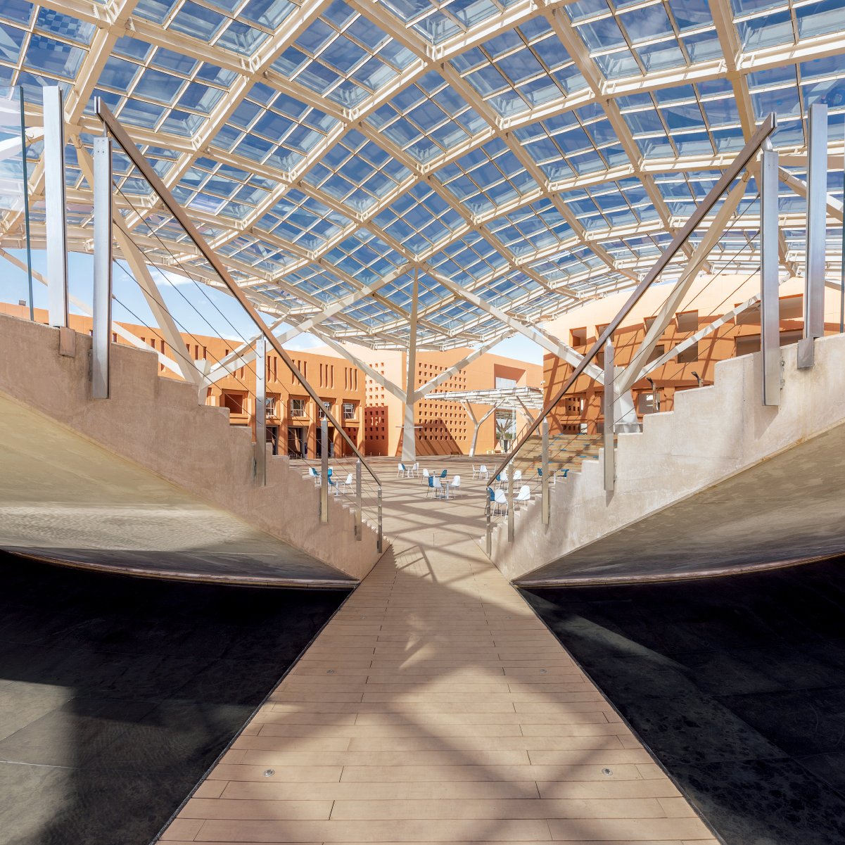 تظهر الصورة تصميمًا معماريًا حديثًا يتميز بسقف زجاجي ضخم يوفر إضاءة طبيعية وفيرة. الأرضية تحت السقف موزعة بطريقة فريدة، حيث تبرز ممرات خشبية وسطحيات مستوية، وتتنوع الأشكال الهندسية بين المنحنيات والزوايا الحادة. يعتبر هذا التصميم تجسيدًا للتوازن بين الطبيعة والتكنولوجيا، مما يعكس أسلوبًا عصريًا ومبتكرًا في البناء.