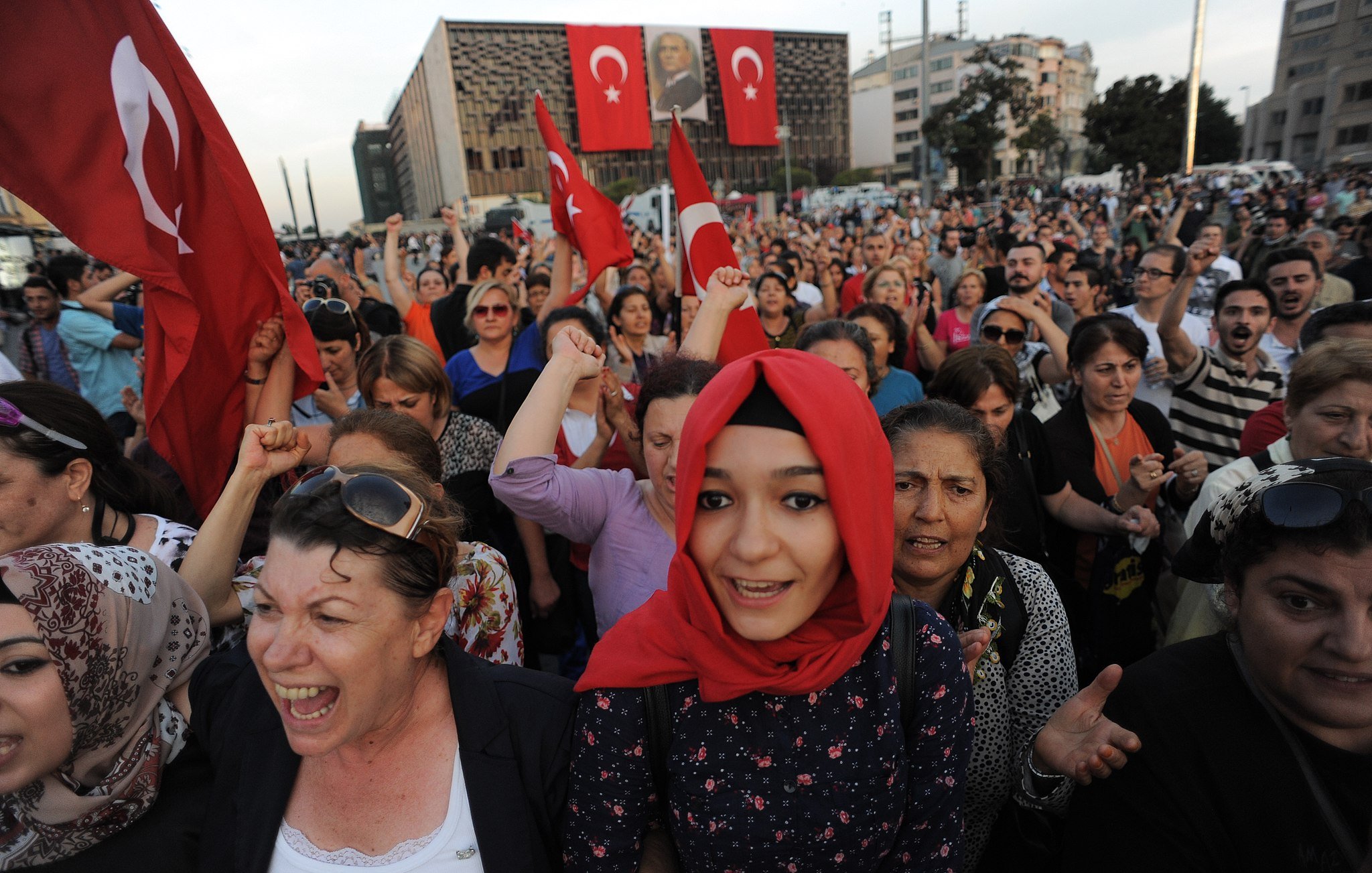 تظهر في الصورة حشود من الناس تجمعوا في مكان واحد، حيث يحمل بعضهم الأعلام التركية. يبدو أن هناك مشاعر حماسية واضحة بين المتظاهرين، حيث يرفعون أصواتهم ويعبرون عن آرائهم. ترتدي بعض النساء الحجاب، بينما تظهر في المقدمة فتاة صغيرة ترتدي غطاء رأس. الخلفية تضم مباني وبعض الصور التي تعكس أجواء الاحتجاج أو الاحتفاء.