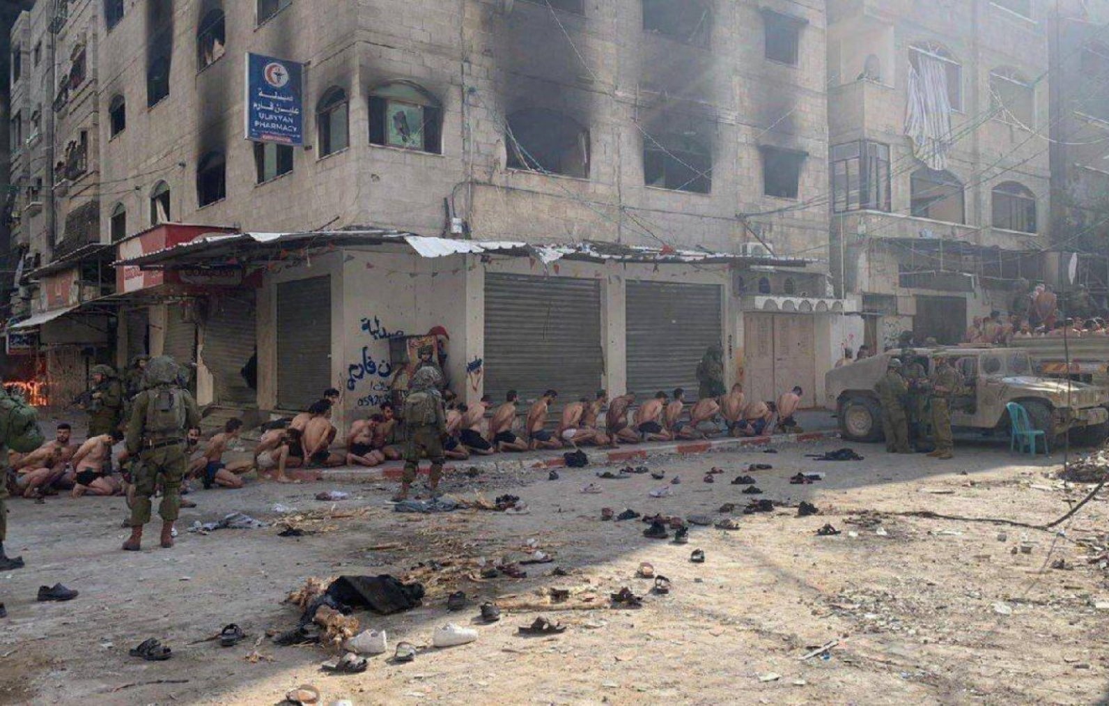 Scène de rue sombre avec des personnes assises et des soldats armés. Atmosphère tendue.