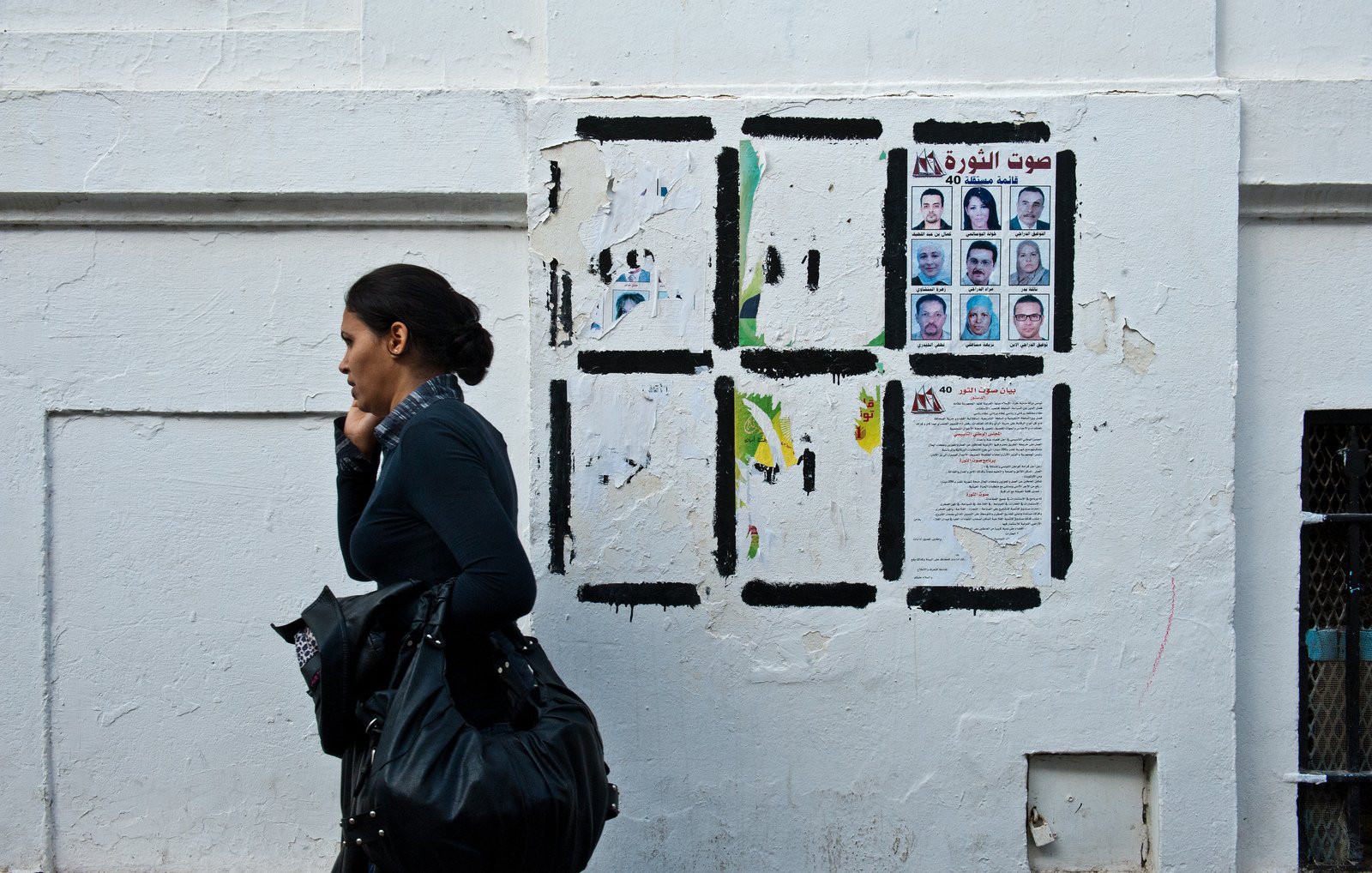 L'image montre une femme marchant le long d'un mur blanc. Elle porte un haut noir et une écharpe. Le mur derrière elle est décoré de divers papiers affichés, y compris des portraits de plusieurs personnes et des textes en arabe. L'atmosphère de la scène semble urbaine et vivante, avec un contraste entre la femme et le mur.