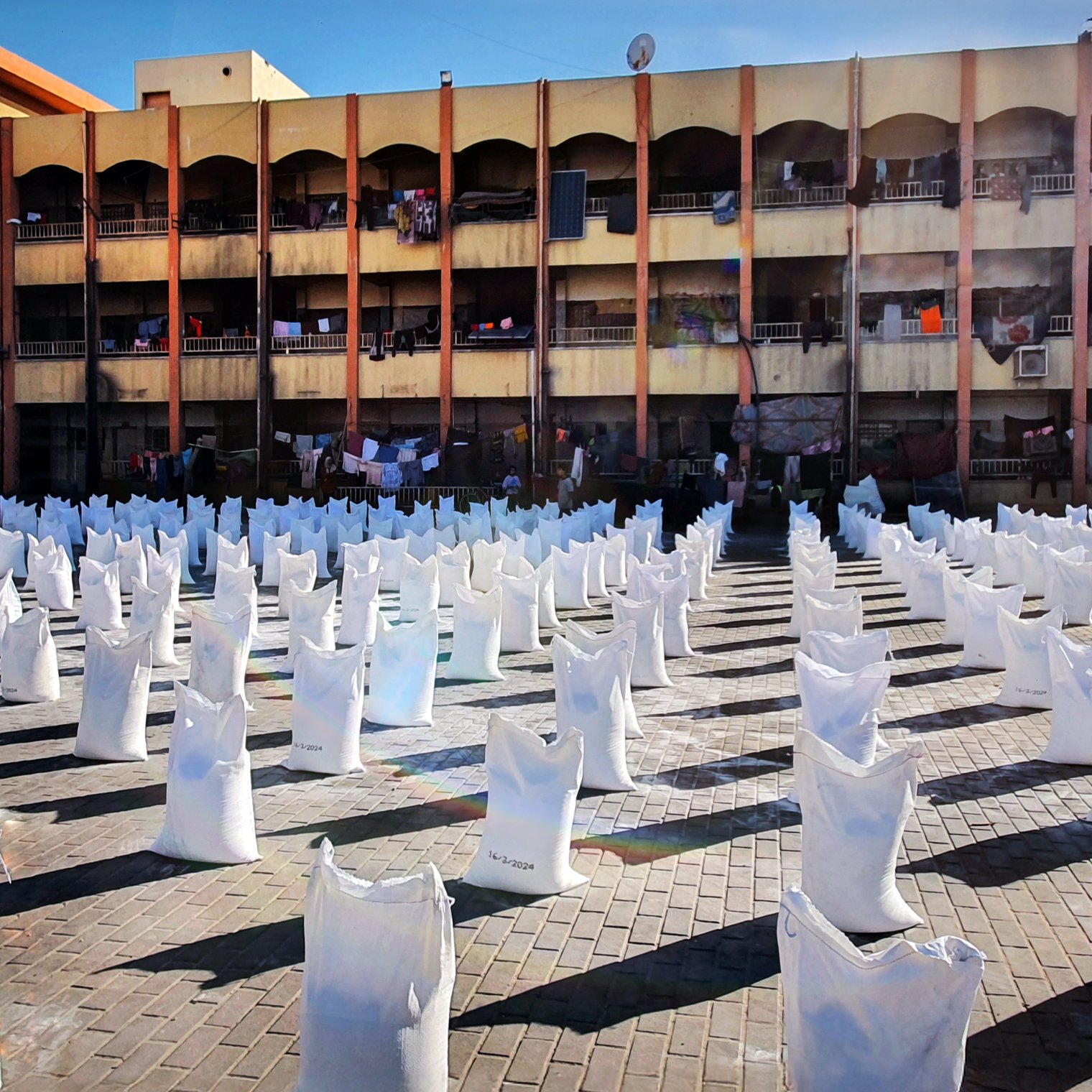 L'image représente un espace ouvert où de nombreux sacs blancs sont disposés en lignes. Ces sacs semblent être remplis de quelque chose, et le sol est constitué de pavés. En arrière-plan, on peut voir un bâtiment à plusieurs étages avec des balcons. Des vêtements sont suspendus à l'extérieur des balcons, suggérant une utilisation résidentielle de cet espace. La lumière du soleil éclaire la scène, créant un contraste entre les sacs blancs et l'environnement.