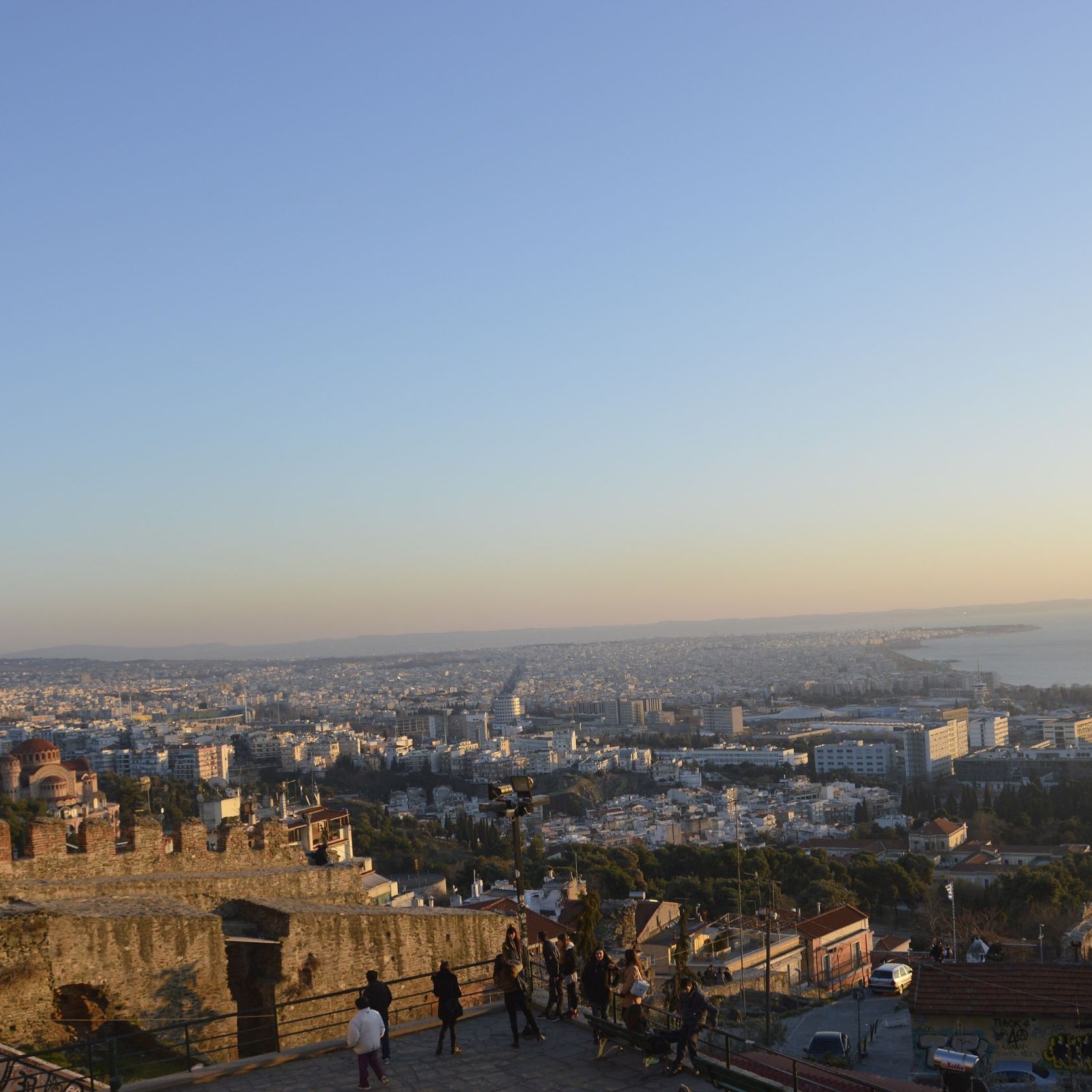 L'image montre une vue panoramique d'une ville au crépuscule. On peut voir un paysage urbain avec de nombreux bâtiments s'étendant jusqu'à l'horizon. À droite, la mer s'étend sur plusieurs kilomètres, et on peut apercevoir quelques bateaux au loin. Au premier plan, on distingue une forteresse ou un château en ruine, avec des murs en pierre. Des personnes se promènent le long d'un chemin, profitant de la vue. La lumière du soir donne une ambiance chaleureuse à la scène.