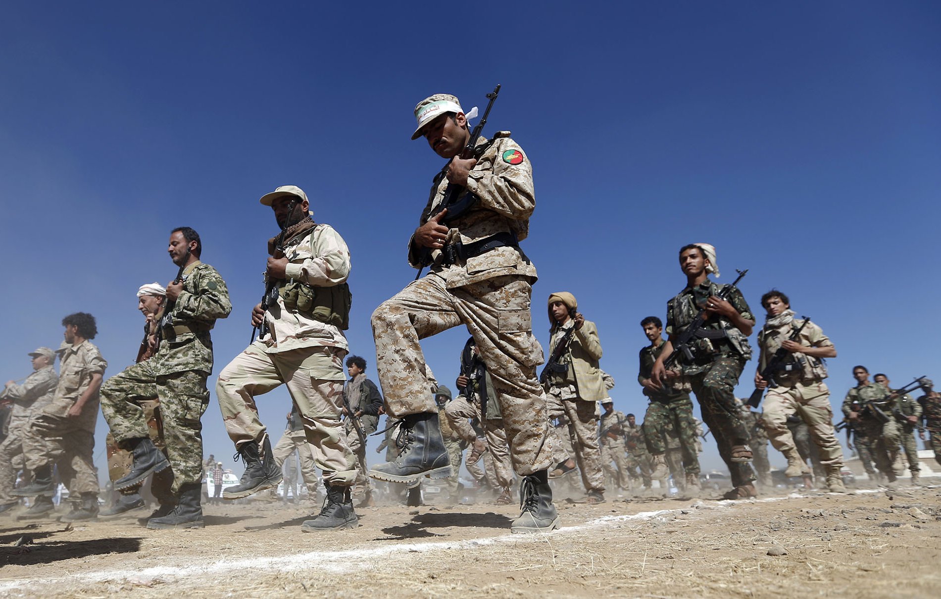 در این تصویر، گروهی از افراد نظامی در حال تمرین یا رژه هستند. آنها لباس نظامی به تن دارند و برخی از آنها سلاح در دست دارند. زمینه تصویر آبی است و به نظر می‌رسد در حال انجام تمرینات نظامی در فضای باز هستند. گرد و غبار در هوا دیده می‌شود که به حرکت آنها تأکید می‌کند. افراد در حال اجرای حرکات هماهنگ و با نظم هستند.