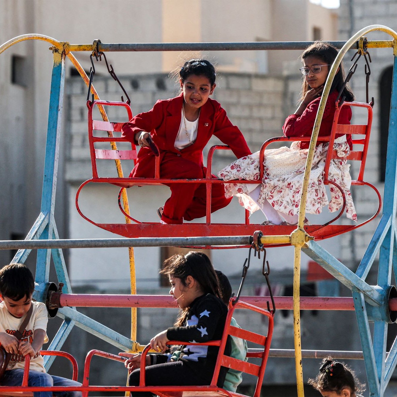 L'immagine mostra un'area giochi all'aperto, con una giostra colorata. Alcuni bambini sono seduti su seggiolini rossi mentre si divertono. Uno dei bambini indossa un completo rosso e sembra divertirsi molto, mentre un'altra bambina, vestita con un abito bianco con fiori, osserva da un'altezza superiore. Gli altri bambini sono anche loro intenti a giocare, creando un'atmosfera di gioia e spensieratezza in un contesto urbano.