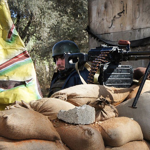 L'image montre un soldat en arrière-plan, vêtu d'un uniforme et portant un casque. Devant lui, il y a une mitrailleuse montée sur un trépied, posée sur un tas de sacs de sable. Le contexte semble être une zone de conflit, avec des éléments de construction en arrière-plan, probablement dans une région touchée par des combats. La scène évoque des mesures de protection et de défense.
