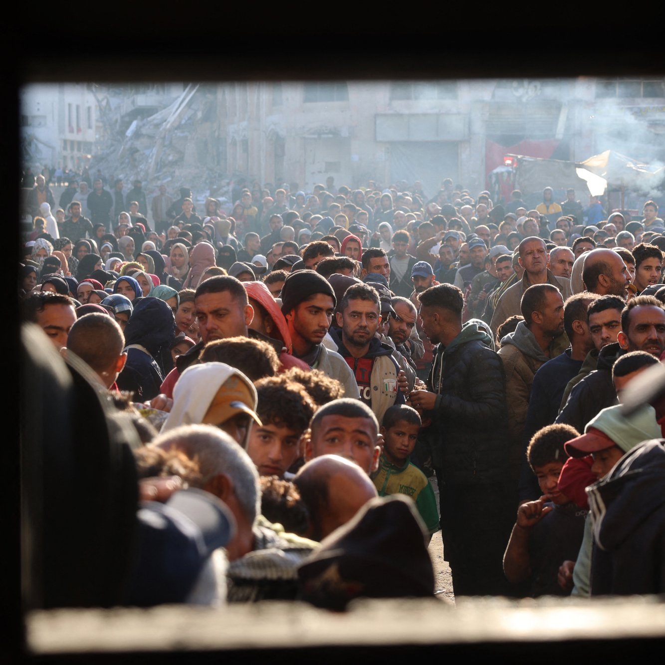 L'immagine ritrae una grande folla di persone in un'area urbana. Si osservano uomini, donne e bambini, tutti concentrati in uno spazio ristretto. L'atmosfera sembra intensa e caotica, con molte persone che si affollano. La luce del sole filtra tra gli edifici, creando un contrasto tra ombre e zone illuminate. La scena potrebbe suggerire un evento significativo o una situazione di emergenza.
