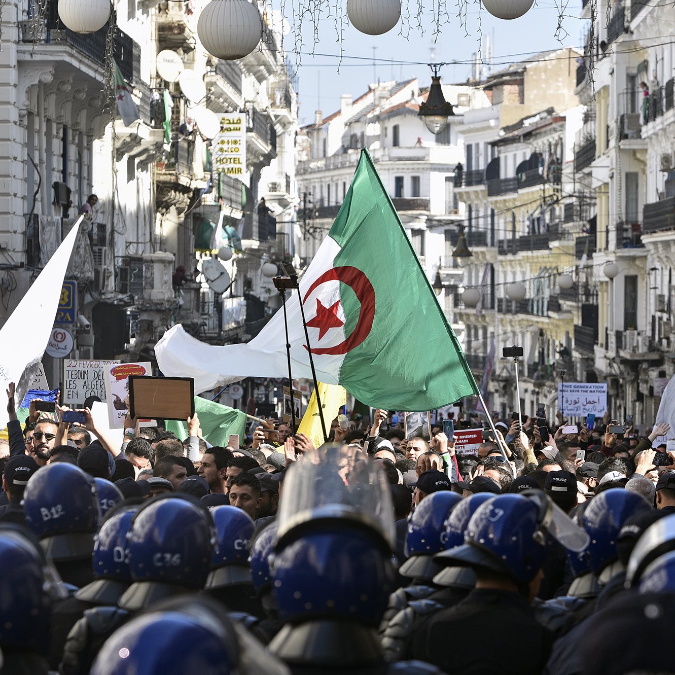 L'image montre une foule rassemblée dans une rue animée, probablement dans une ville algérienne, en période de manifestation. Des personnes portent des drapeaux algériens et tiennent des pancartes, tandis qu'un groupe de policiers en uniforme, portant des casques, se tient en ligne devant eux. Les bâtiments alentours sont typiques d'une architecture urbaine, ornés de décorations. L'atmosphère semble être celle d'une démonstration populaire, avec un fort engagement civique visible parmi les manifestants.