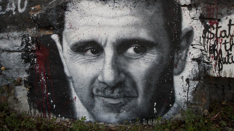 L'image montre un graffiti représentant un visage en noir et blanc sur un mur en pierre. Les traits du visage sont bien définis, avec un regard intense et expressif. Le fond est partiellement recouvert de peinture, et on peut voir des éclaboussures de couleur rouge, ajoutant une dimension dramatique à l'œuvre. Ce type d'art urbain est souvent utilisé pour faire passer des messages ou des commentaires sociaux.