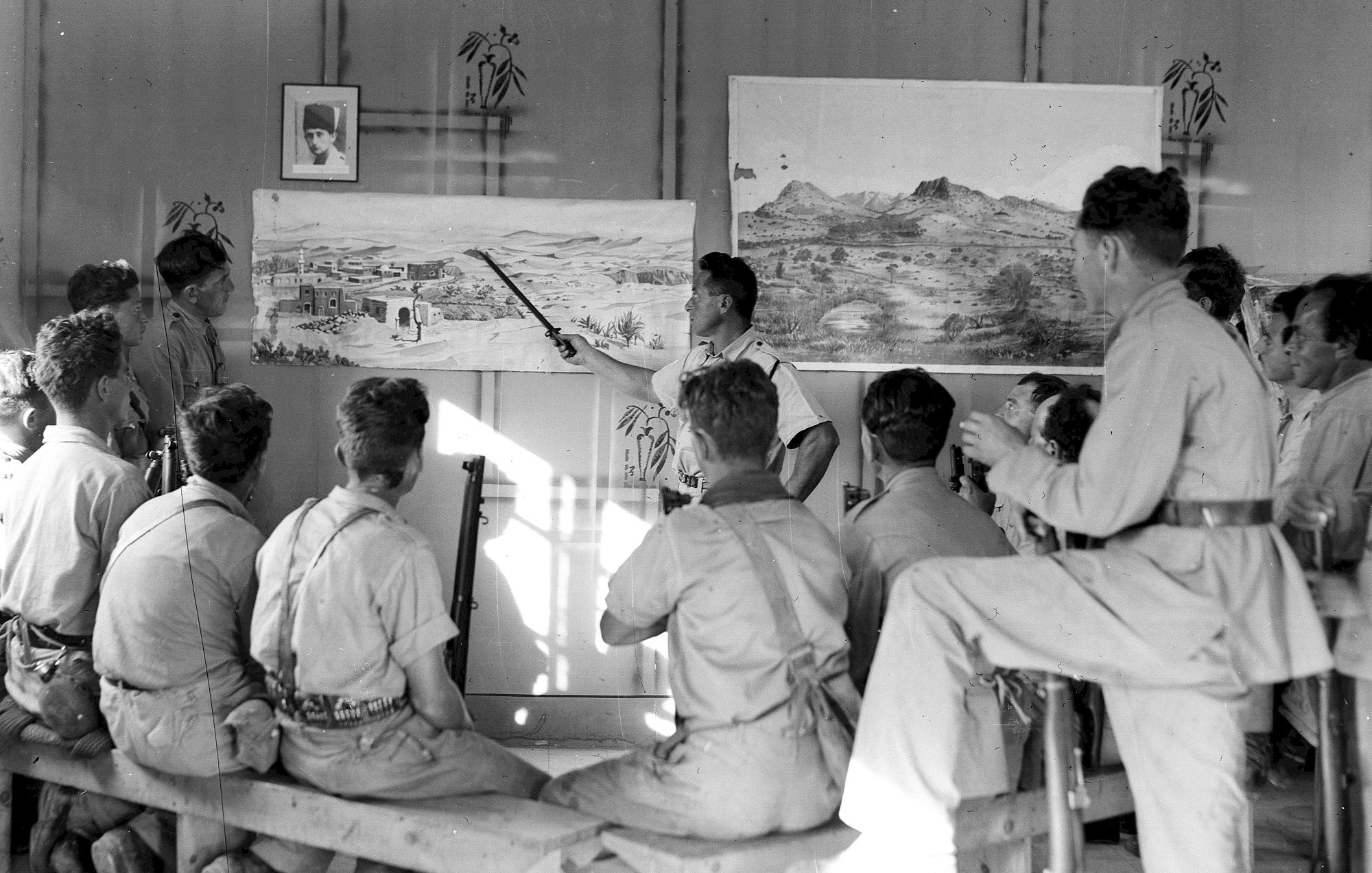 L'image montre un groupe de personnes assises sur des bancs, attentives à un homme qui semble donner une présentation devant des tableaux accrochés au mur. Les tableaux illustrent des paysages, l'un représentant probablement un village ou une scène de la nature. L'environnement est dépouillé, avec des murs simples et une lumière naturelle qui pénètre à travers une fenêtre. Les participants, majoritairement en vêtements uniformes, semblent captivés par l'explication de l'intervenant. Il y a aussi un portrait encadré sur le mur, ajoutant un élément de contexte à la scène.