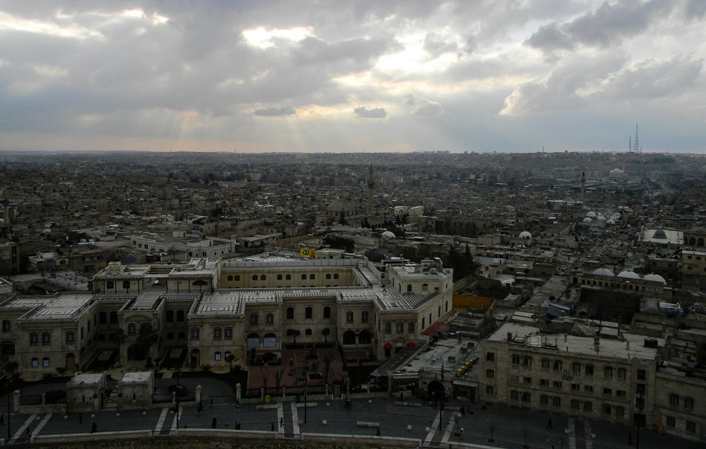 L'image montre une vue panoramique d'une ville, probablement une ancienne ville, avec des bâtiments en pierre grise et une architecture typique du Moyen-Orient. Le ciel est nuageux, avec des rayons de lumière qui percent à travers les nuages, créant une atmosphère dramatique. À l'arrière-plan, on peut apercevoir d'autres structures urbaines et des mosquées, indiquant une richesse historique. L'ambiance semble à la fois paisible et chargée de mélancolie.