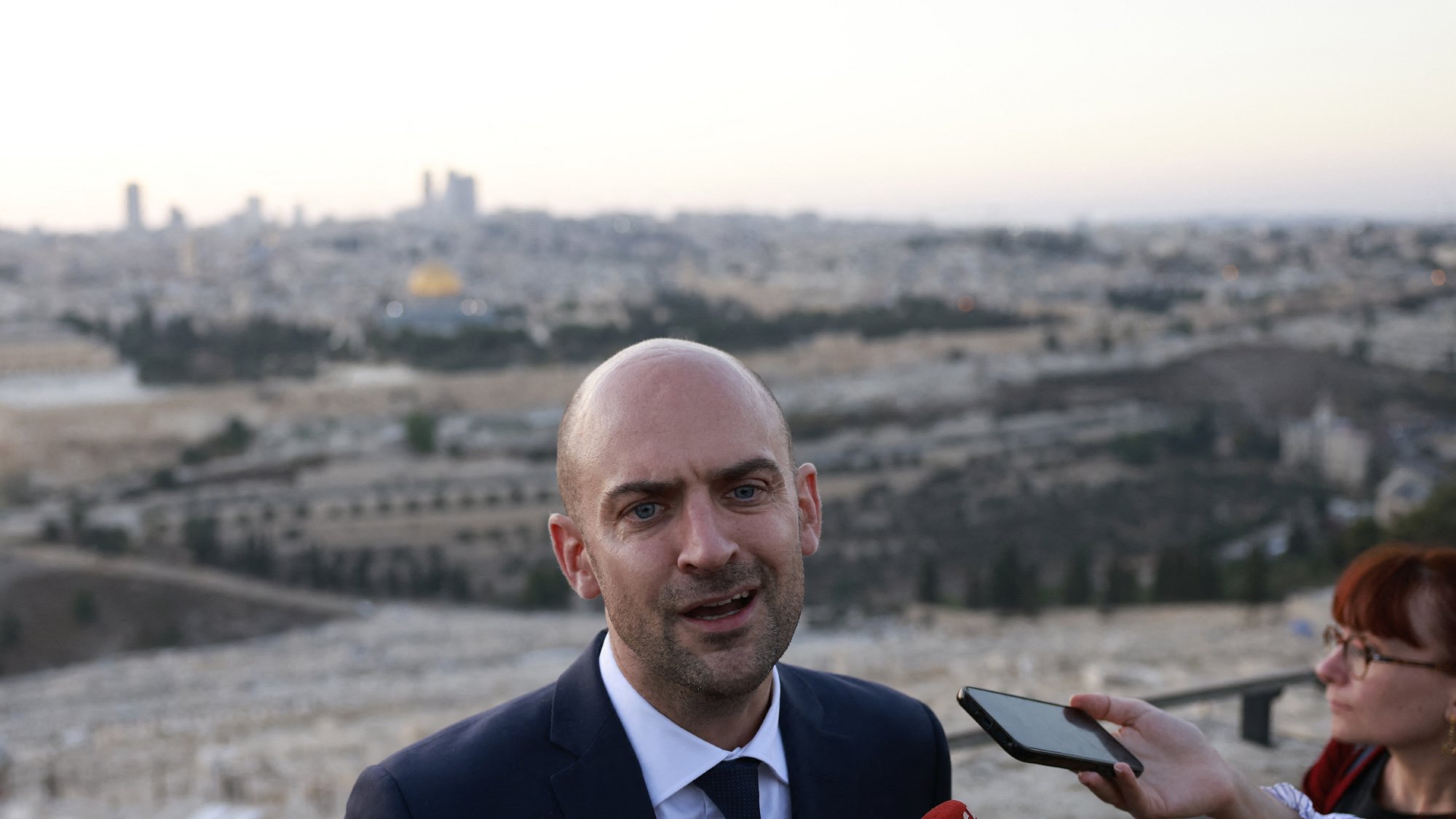 در این تصویر، یک مرد با سر طاس و کت و شلوار تیره در حال صحبت به دوربین و خبرنگاران است. او در یک مکان آزاد ایستاده و پشتش چشم‌انداز شهر قابل مشاهده است. درختان و ساختمان‌ها در دوردست دیده می‌شوند، و به نظر می‌رسد که این تصویر در یک زمان غروب یا صبح زود گرفته شده است. خبرنگاری نیز در سمت راست او قرار دارد و در حال ثبت‌کردن یا پرسش کردن از اوست.