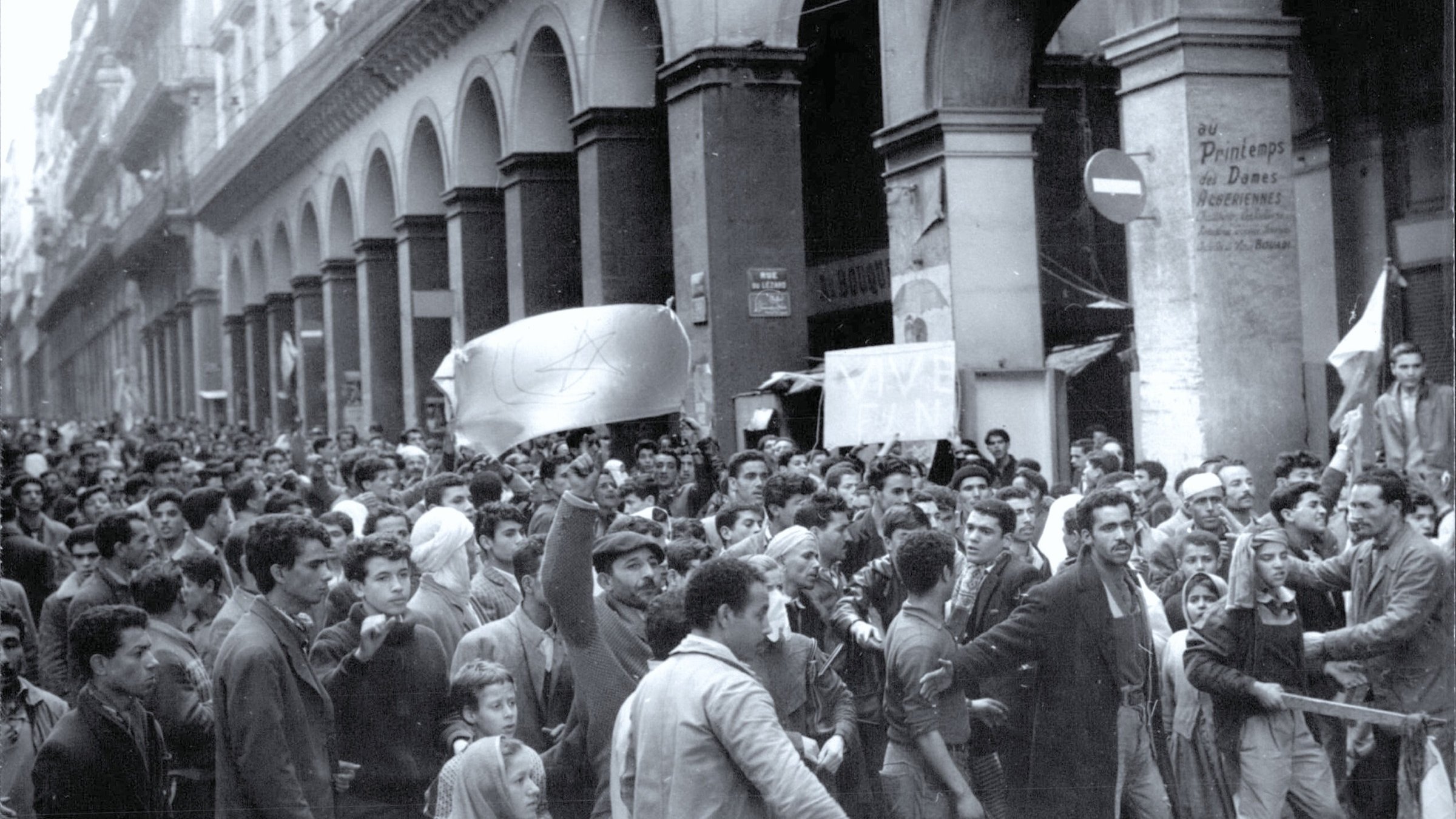 L'image montre une grande manifestation dans une rue animée, où une foule dense est rassemblée. Les manifestants semblent exprimer des revendications, brandissant des drapeaux et des pancartes. L'architecture environnante est typiquement urbaine, avec de grands arcs et des bâtiments anciens. L'atmosphère générale semble passionnée et engagée, reflétant un fort esprit de protestation.