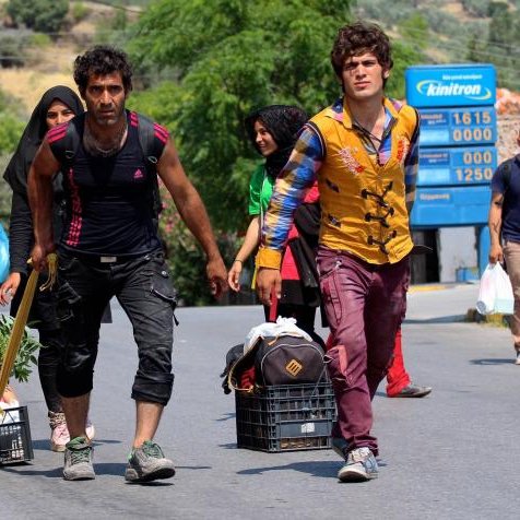 L'image montre un groupe de personnes se déplaçant le long d'une route. Il y a des enfants et des adultes, certains portent des vêtements colorés. Deux hommes avancent avec des paniers à roulettes, contenant probablement des biens ou de la nourriture. L'un d'eux tient une plante, tandis que l'autre semble porter une baguette ou un long objet. On peut voir des maisons et des arbres en arrière-plan, ajoutant une touche de nature à la scène. L'ambiance générale évoque un déplacement ou un voyage.