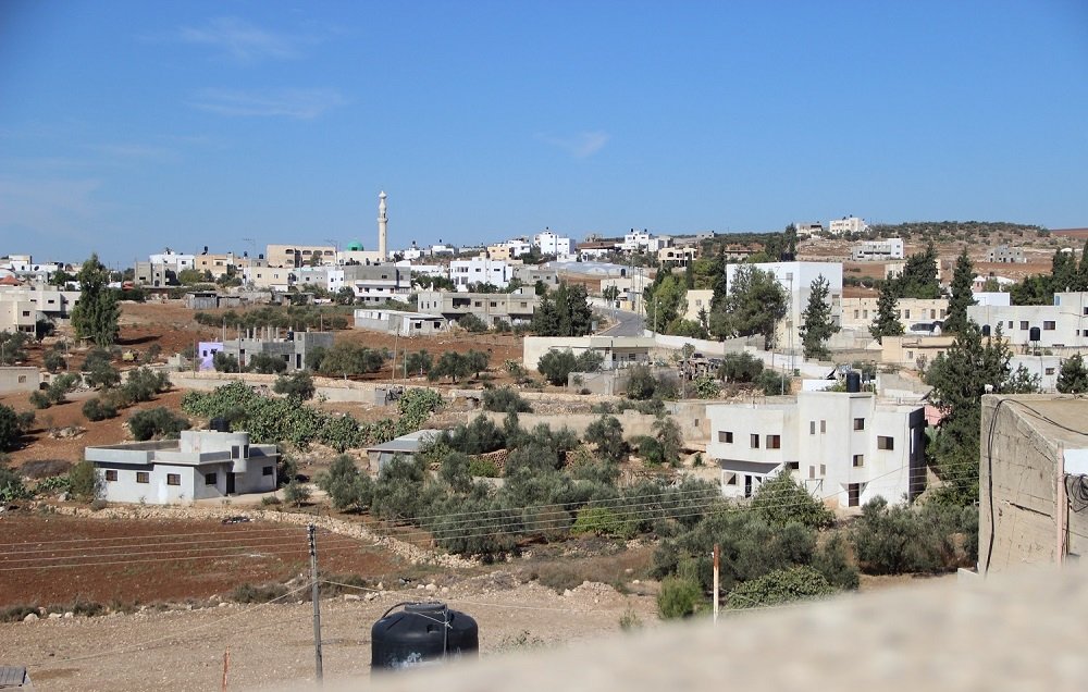 L'image montre un paysage urbain de type rural, avec des maisons de couleur claire disséminées sur une colline. On peut apercevoir des arbres et des champs, ainsi qu'un minaret se dressant dans le fond, ce qui pourrait indiquer la présence d'une mosquée. Le ciel est dégagé et d'un bleu vif, ce qui donne à la scène une atmosphère ensoleillée. Les infrastructures semblent simples et typiques des zones périphériques.