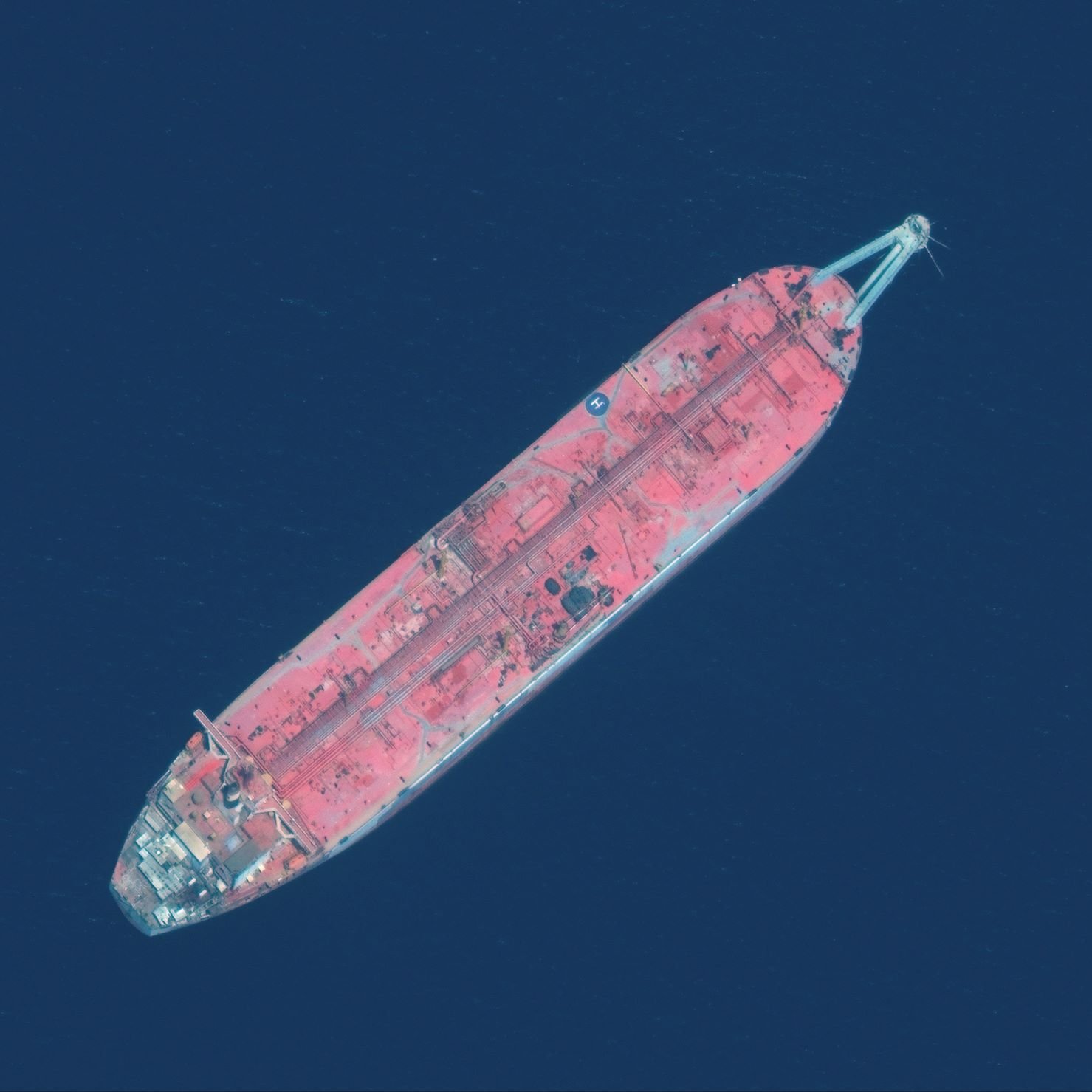 L'image montre un grand navire de transport de pétrole, vu du dessus. Le navire a une coque de couleur rouge, avec des détails visibles sur le pont. Il flotte sur une surface d'eau bleue profonde, ce qui donne un fort contraste entre le navire et son environnement. Les structures sur le pont semblent usées, suggérant un certain âge ou une exposition aux éléments marins.