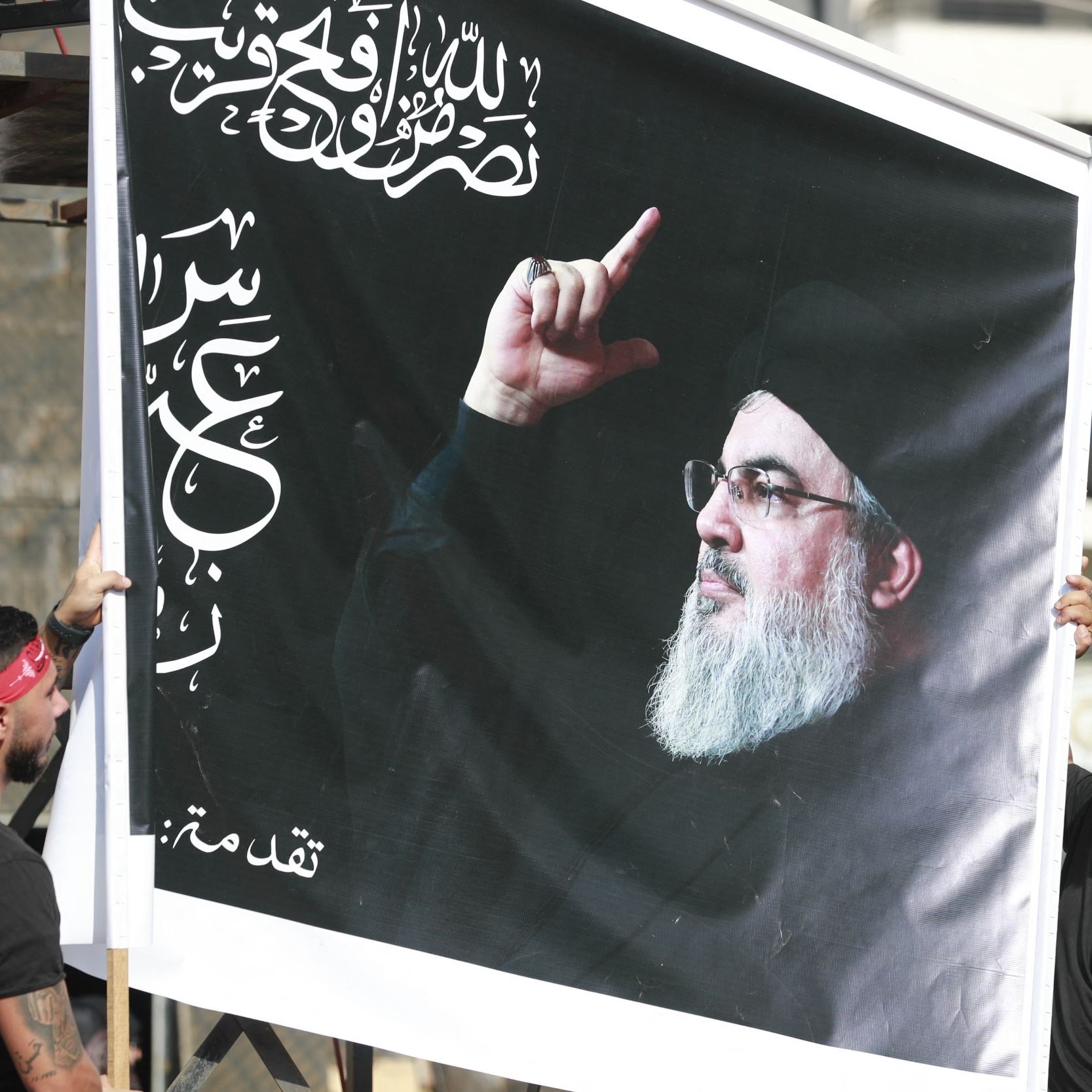 L'immagine mostra due uomini che sorreggono un grande striscione. Sullo striscione è rappresentato il volto di un uomo con un gesto della mano che sembra indicare qualcosa. Il contesto suggerisce una celebrazione o una manifestazione, con i partecipanti che indossano fasce rosse. Lo sfondo è urbanizzato, con edifici visibili.