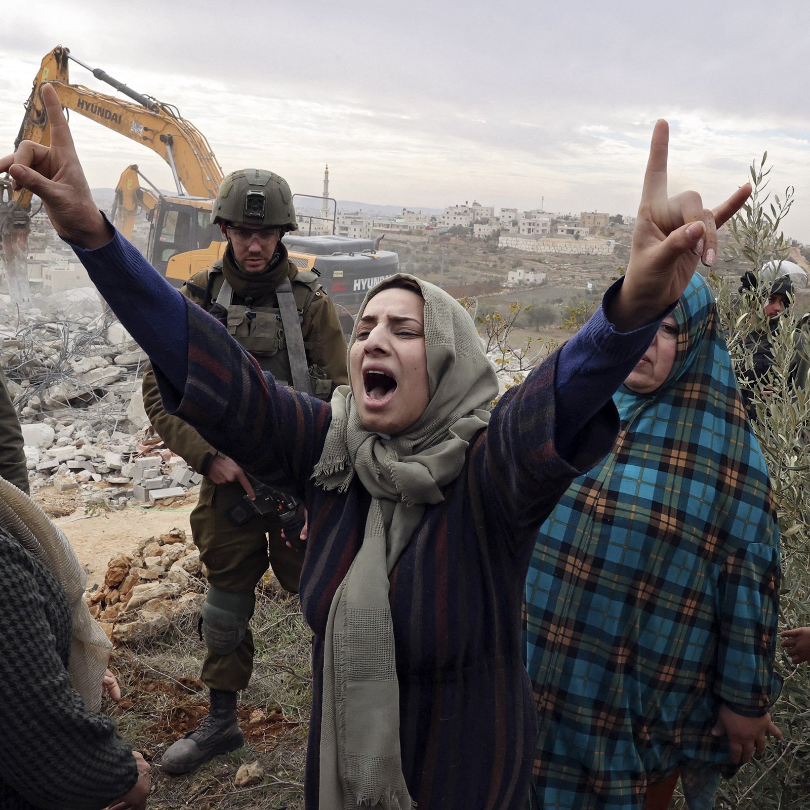 L'image montre une scène intense où une femme exprime sa colère et sa frustration, levant les bras en signe de protestation. Elle est entourée de plusieurs personnes, dont certaines ont l'air préoccupées ou attristées. En arrière-plan, on aperçoit des forces de l'ordre et un engin de construction qui semble démolir une structure. L'atmosphère dégagée par l'image est chargée d'émotion, suggérant des tensions et des luttes en cours.